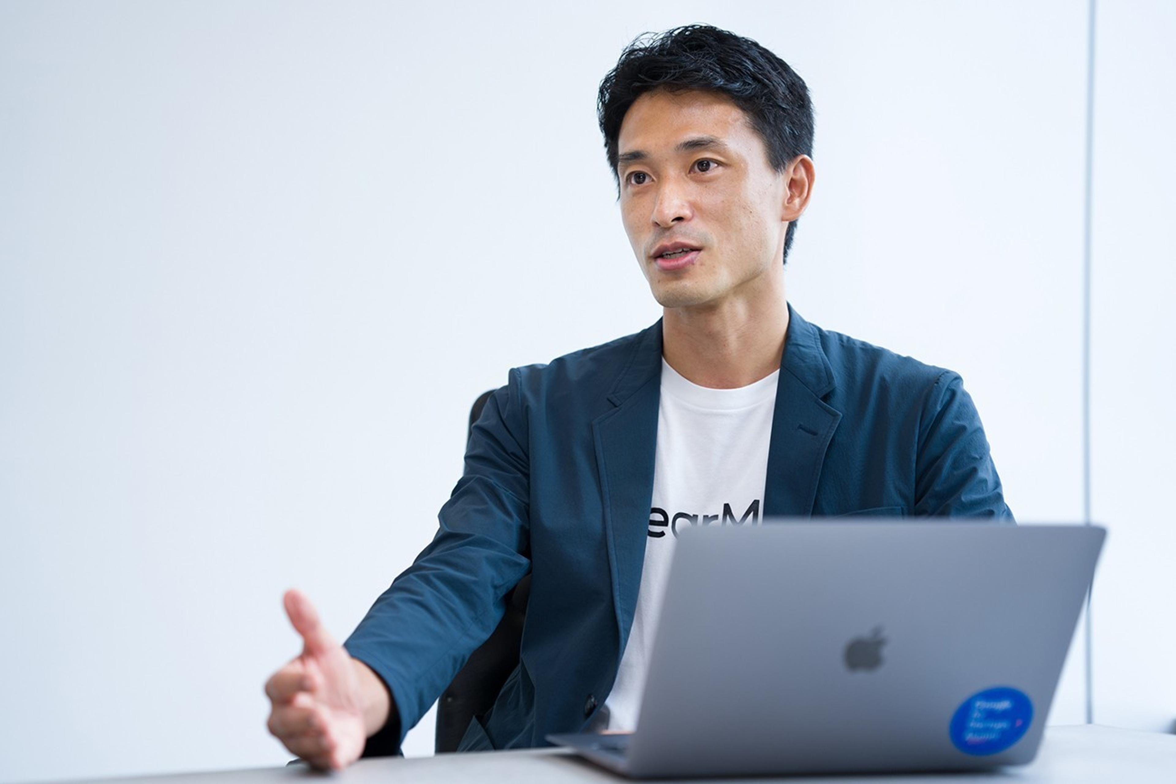 ビジネススーツを着た男性が、ノートパソコンの前で話している様子。背景は白い壁で、シンプルなオフィス環境。