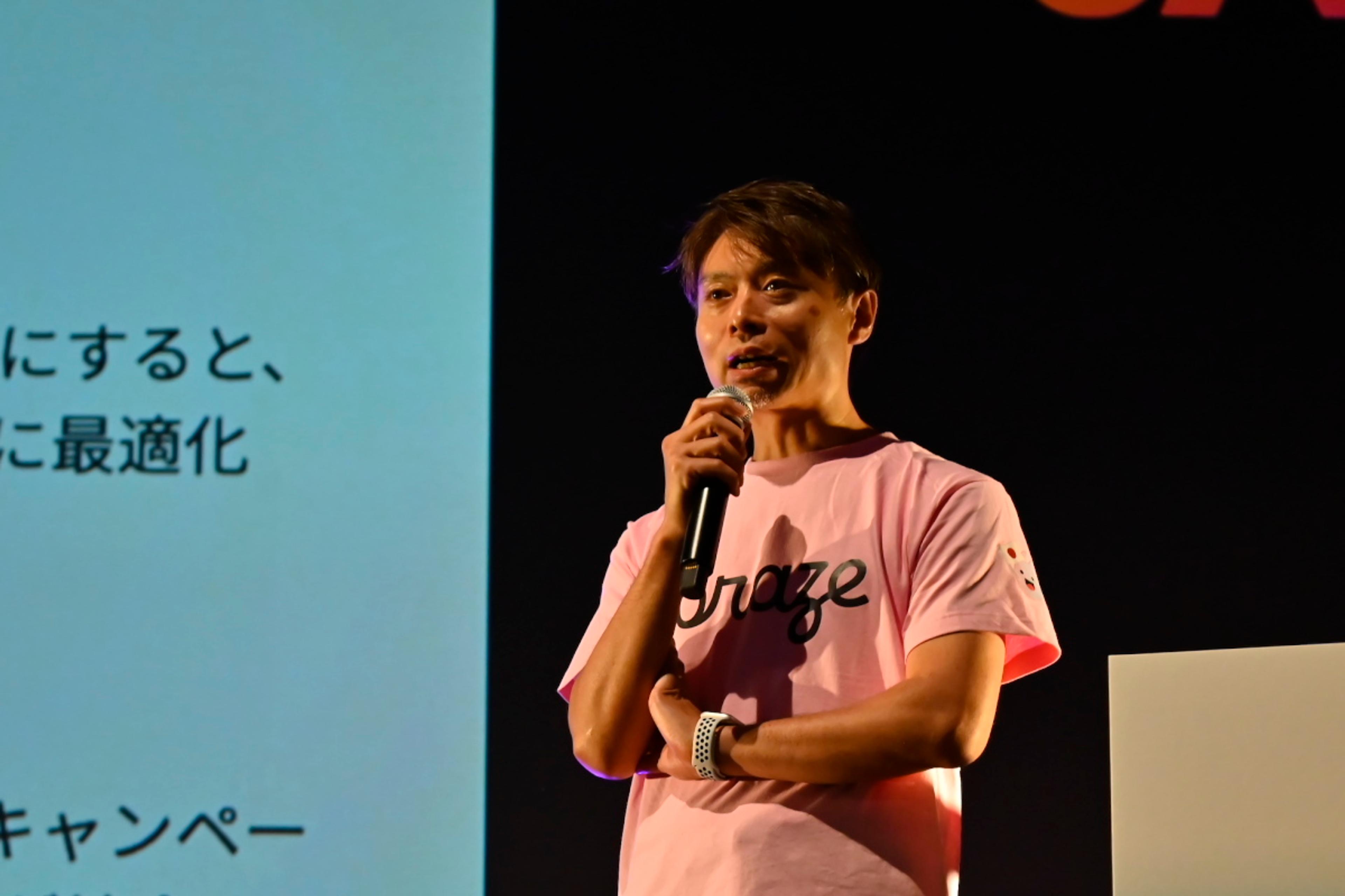 ピンクのTシャツを着た男性がマイクを持って話しています。背景にはプレゼンテーションのスライドがあります。