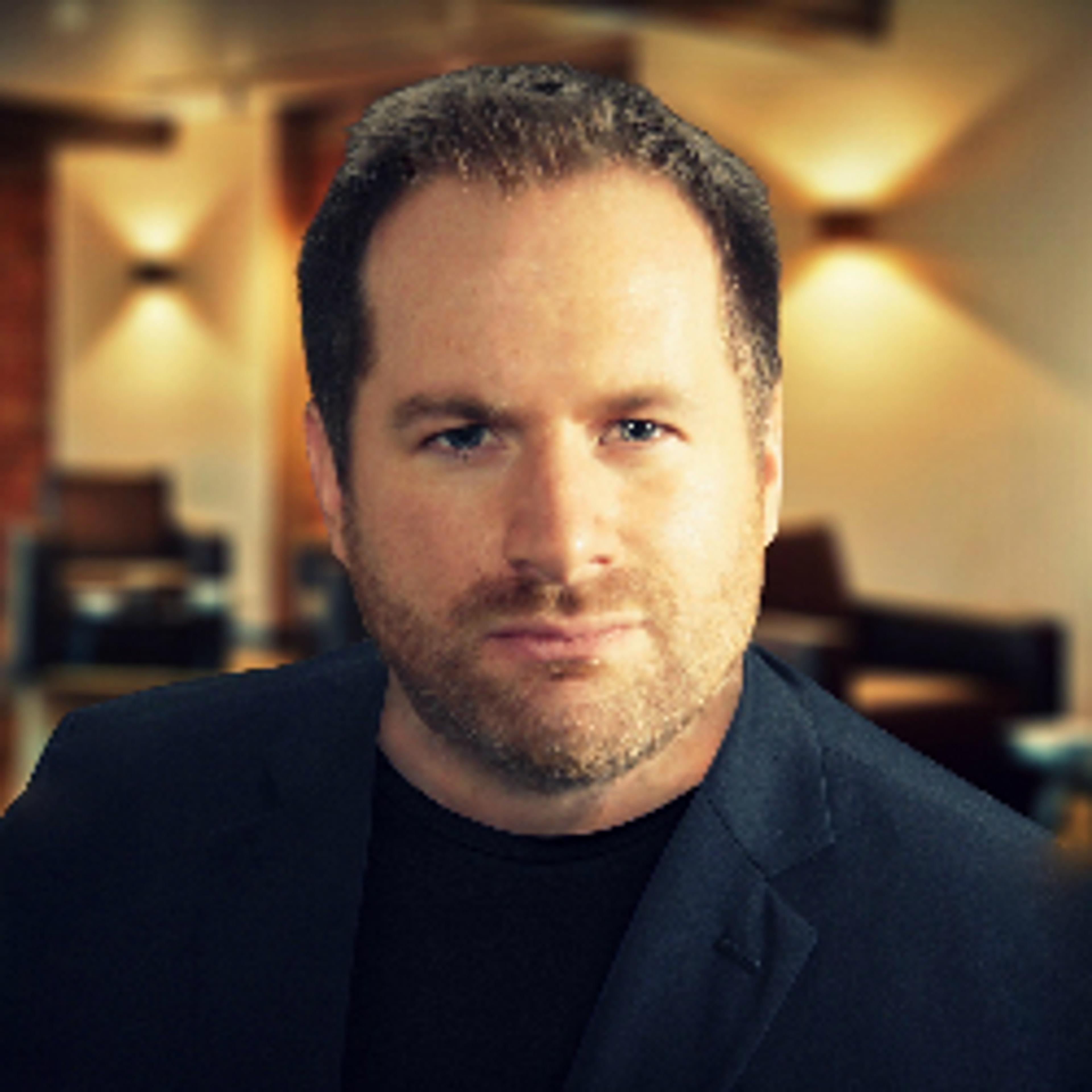 A man with short hair and a beard is wearing a black blazer, looking directly at the camera. The background features a softly lit interior space.