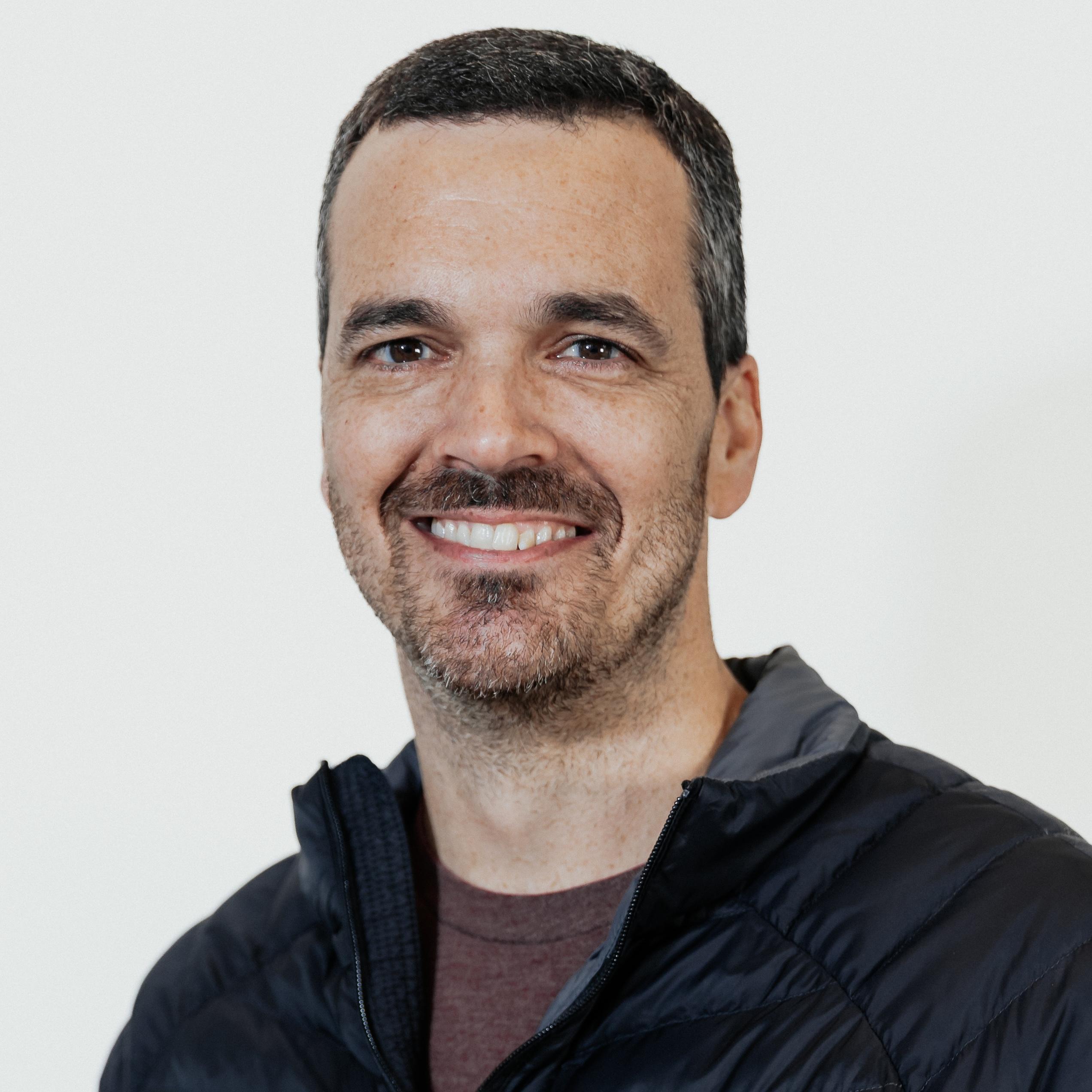 Headshot of a smiling man with short dark hair and a trimmed beard, wearing a black jacket against a light background.