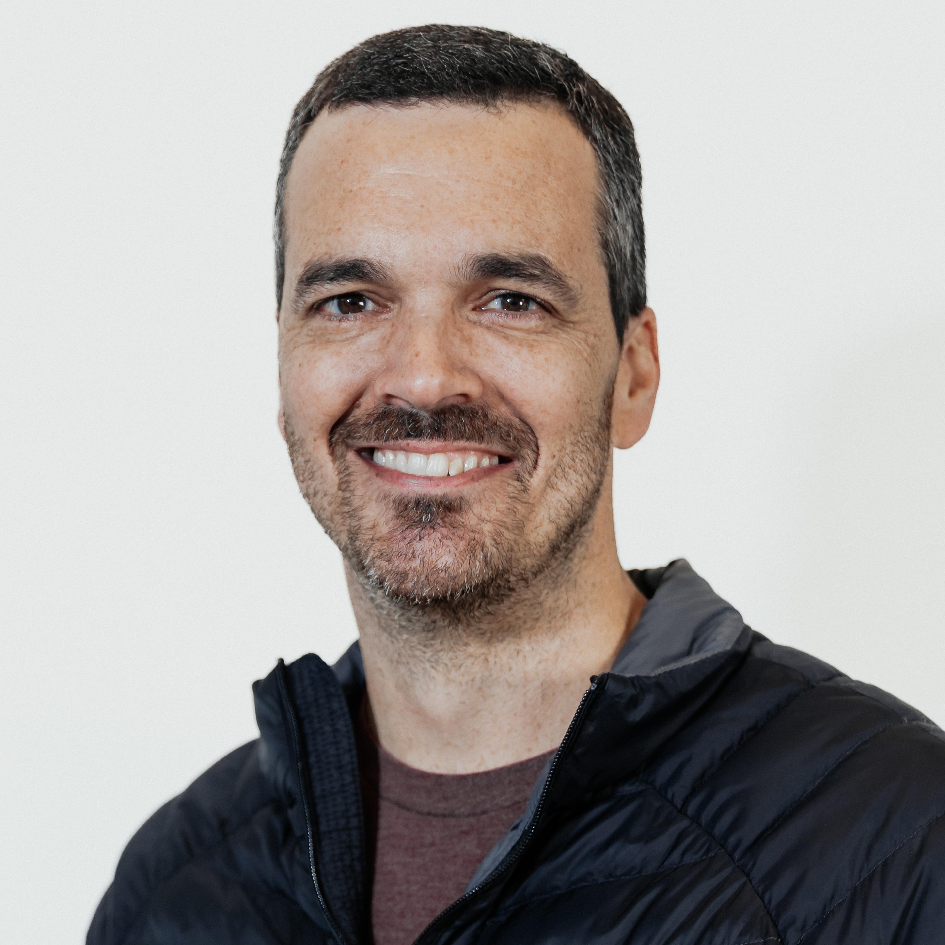 Headshot of a smiling man with short dark hair and a trimmed beard, wearing a black jacket against a light background.