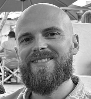 Black-and-white close-up portrait of a smiling bearded person with a shaved head, looking at the camera in an outdoor setting.
