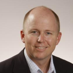 A smiling man with a bald head and short hair, wearing a dark blazer over a light checkered shirt. The background is a soft gradient.