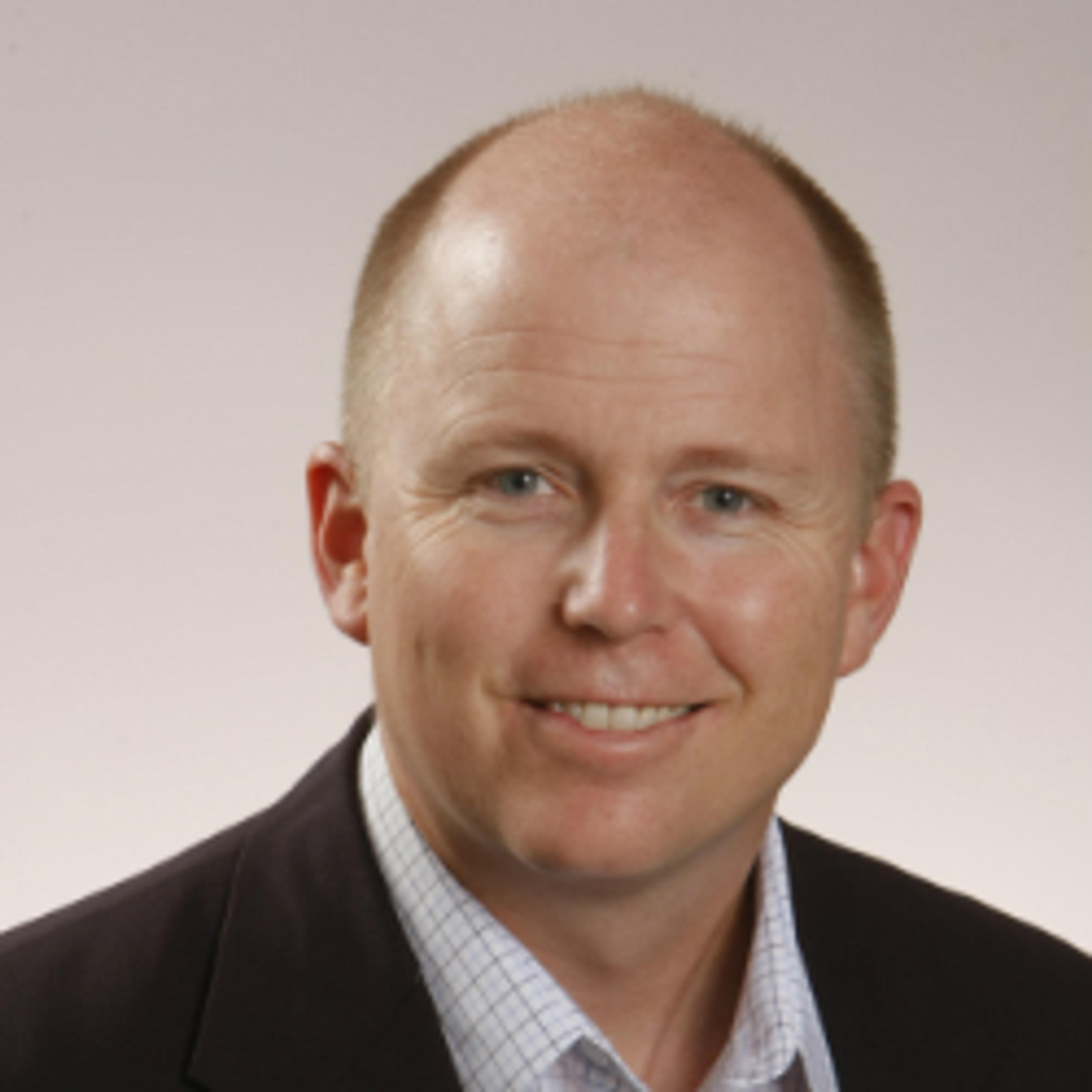 A smiling man with a bald head and short hair, wearing a dark blazer over a light checkered shirt. The background is a soft gradient.