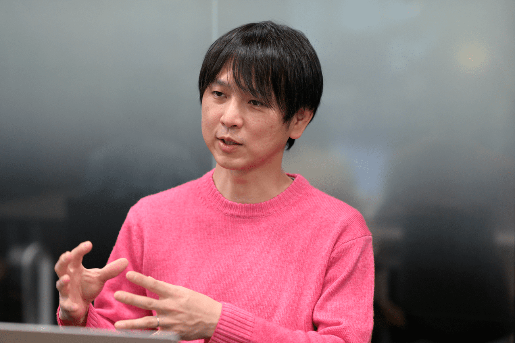 ピンクのセーターを着た男性が話している様子。背景はぼやけたオフィスの風景。