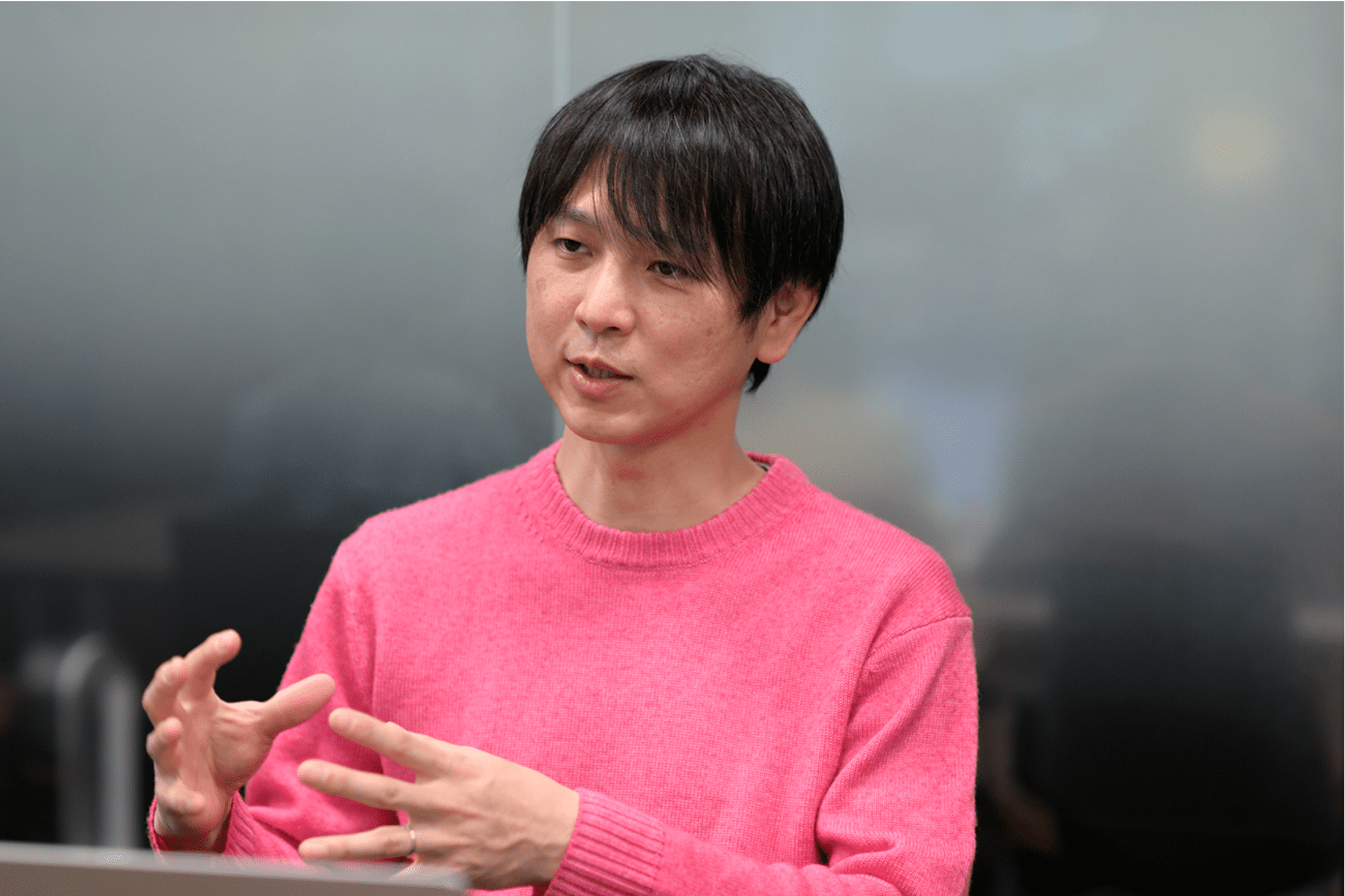 ピンクのセーターを着た男性が話している様子。背景はぼやけたオフィスの風景。