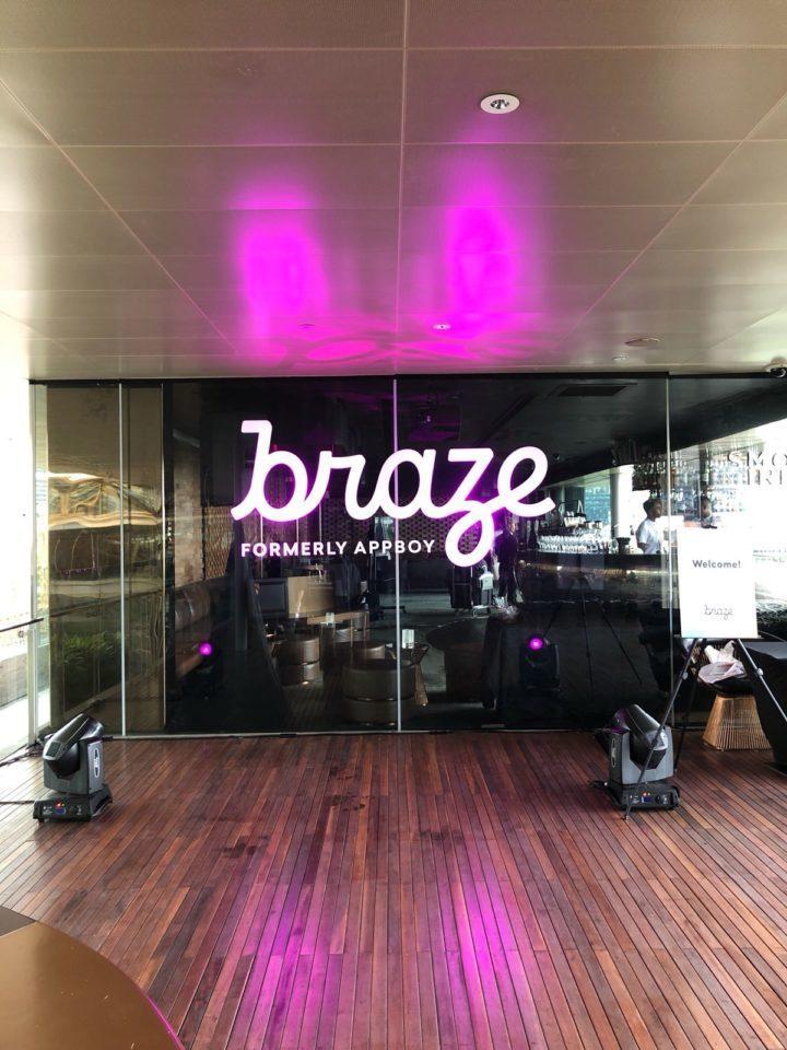 A glass wall displays the logo for Braze, formerly known as Appboy, with purple lighting. The floor is wooden, and there are speakers on either side.