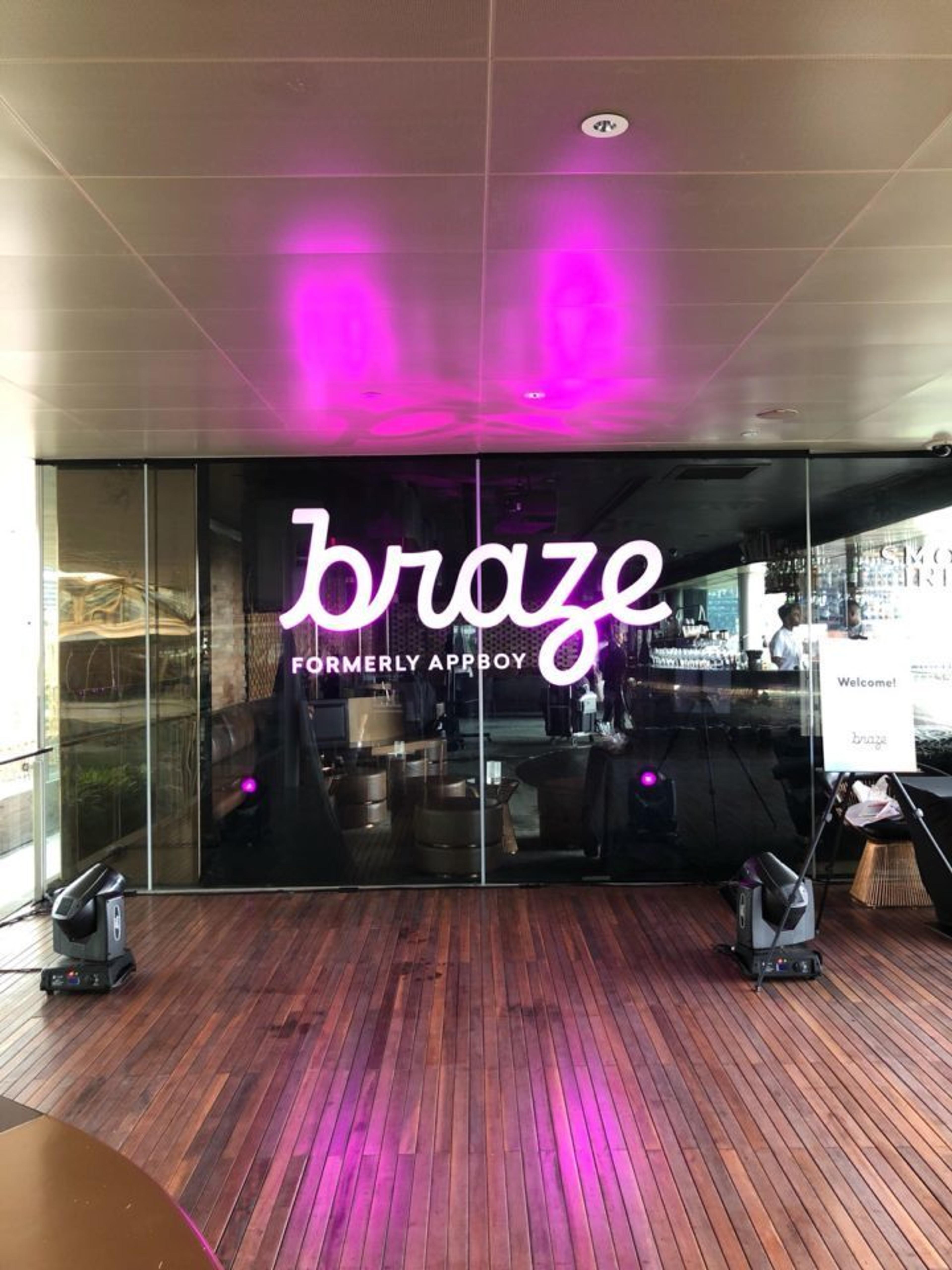 A glass wall displays the logo for Braze, formerly known as Appboy, with purple lighting. The floor is wooden, and there are speakers on either side.