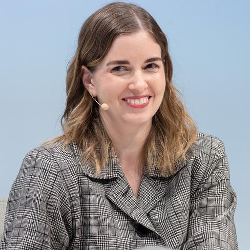 A woman with shoulder-length brown hair smiles while wearing a checkered blazer and a microphone headset. She is seated against a light blue background.