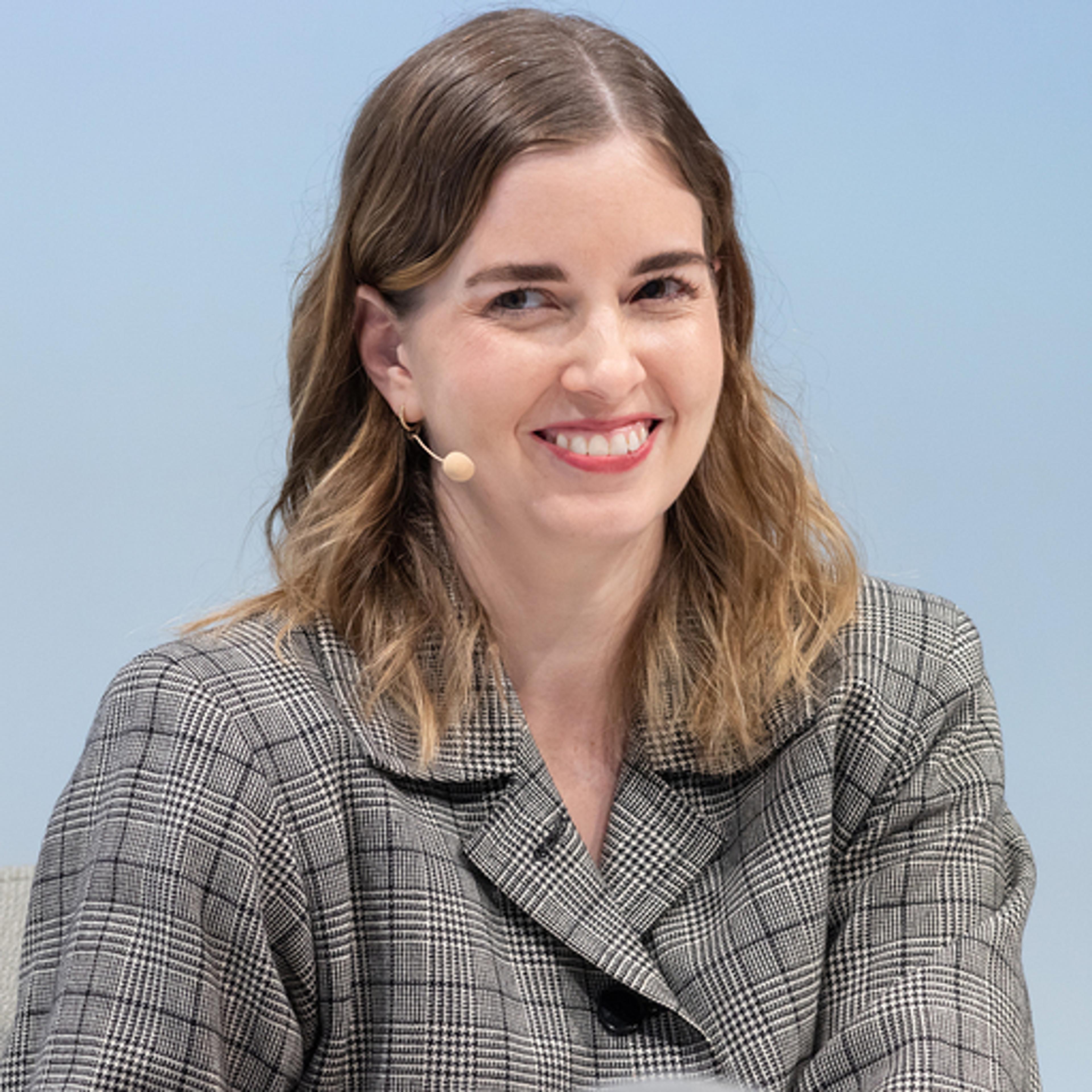 A woman with shoulder-length brown hair smiles while wearing a checkered blazer and a microphone headset. She is seated against a light blue background.