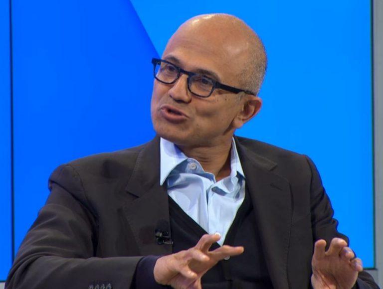 A man with glasses and a bald head is speaking while gesturing with his hands. He is wearing a black suit over a light blue shirt.