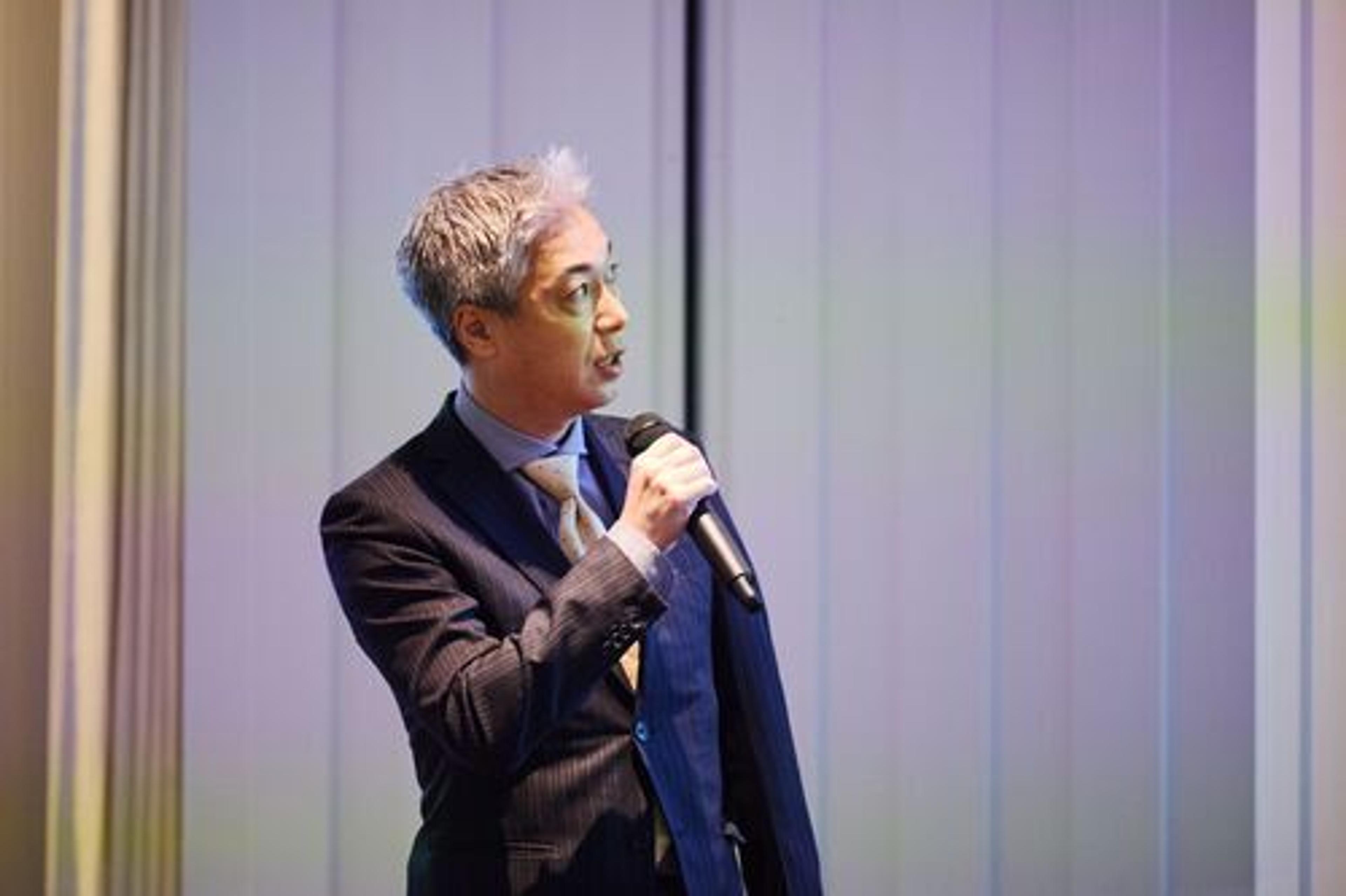 スーツを着た男性がマイクを持って話している。背景は淡い色合いの照明が当たっている。