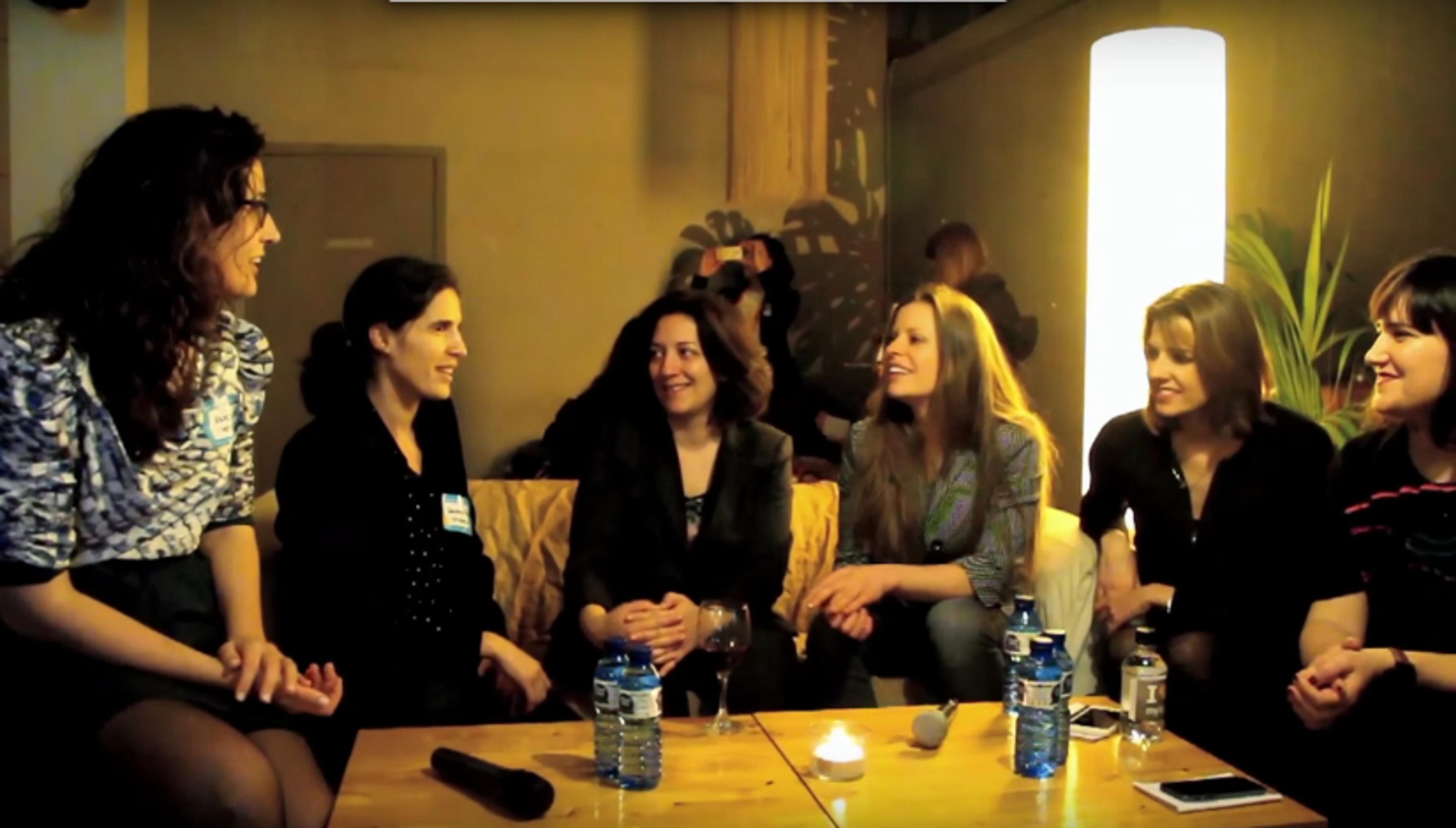 A group of six women are sitting together in a cozy setting, smiling and engaging in conversation. There is a warm light and a small table with water bottles and a candle.