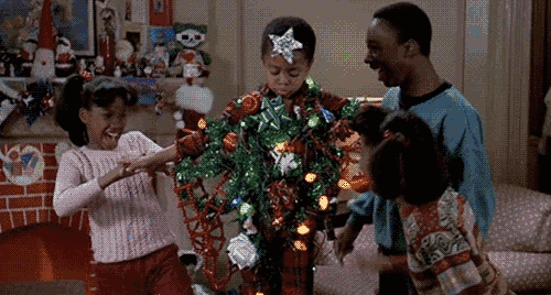 A boy is playfully wrapped in Christmas lights and decorations while three friends laugh and interact around him. The scene is festive and joyful, set in a decorated living room.