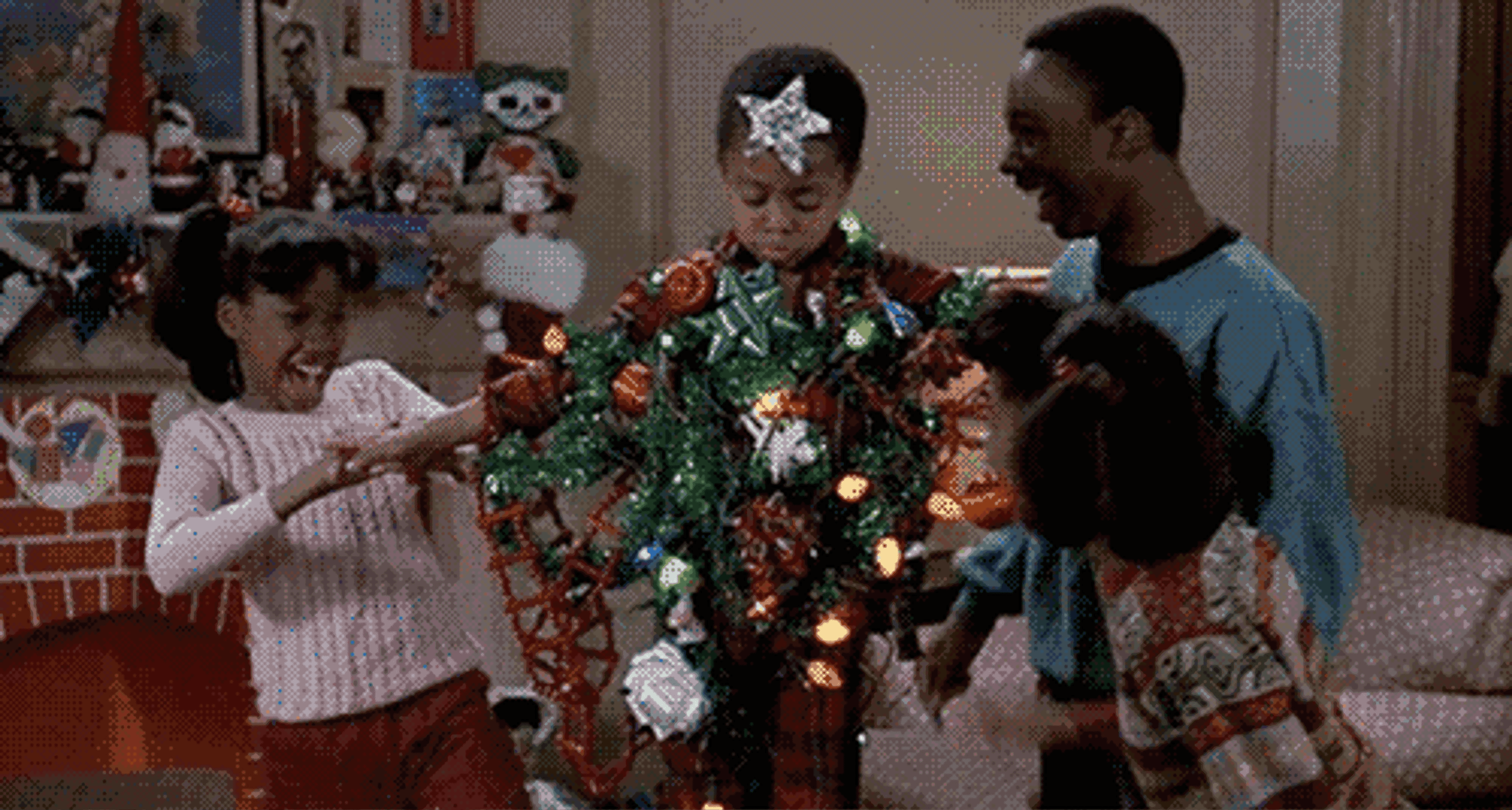 A boy is playfully wrapped in Christmas lights and decorations while three friends laugh and interact around him. The scene is festive and joyful, set in a decorated living room.