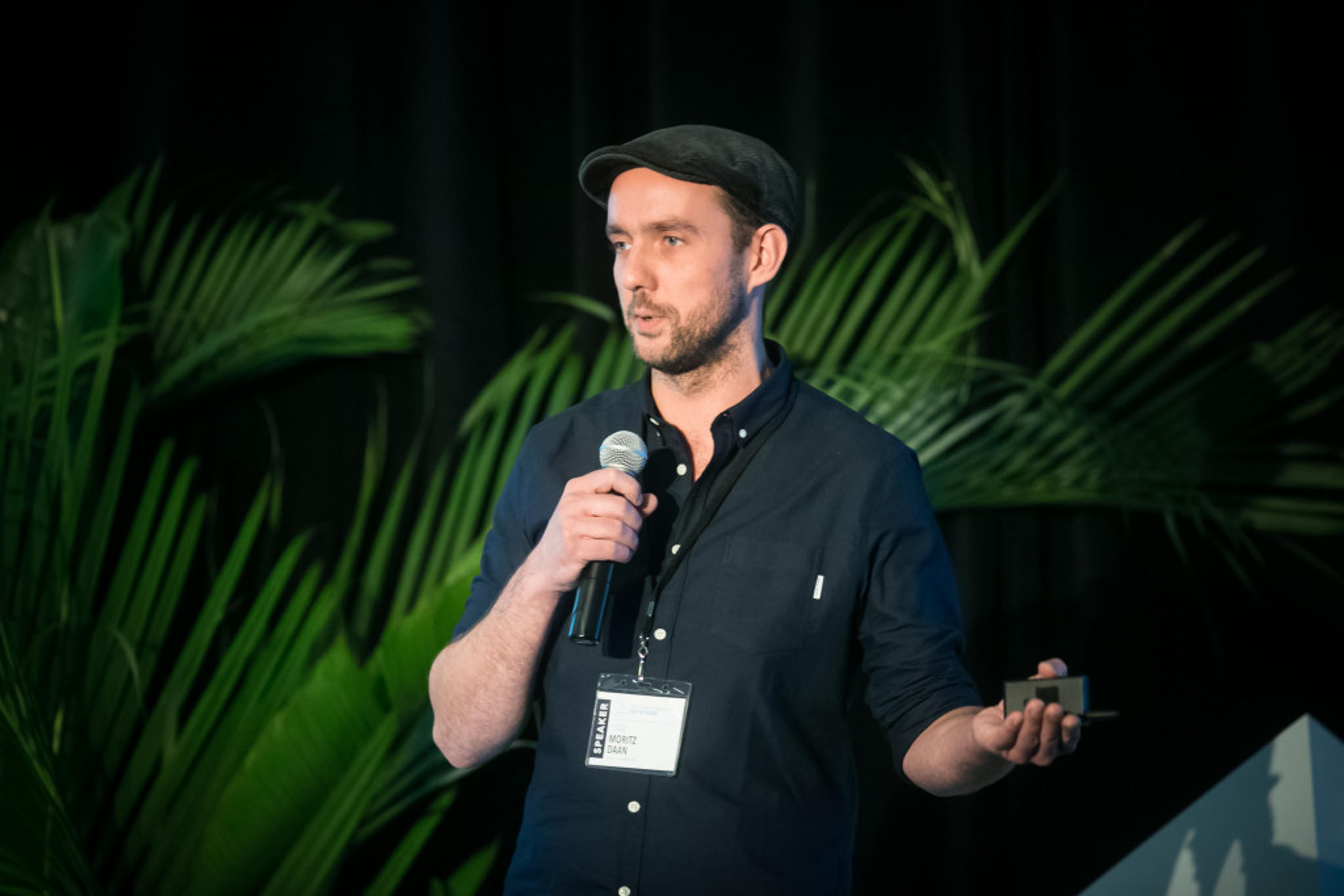 A man in a black shirt and cap speaks into a microphone while holding a small device, with green plants in the background.