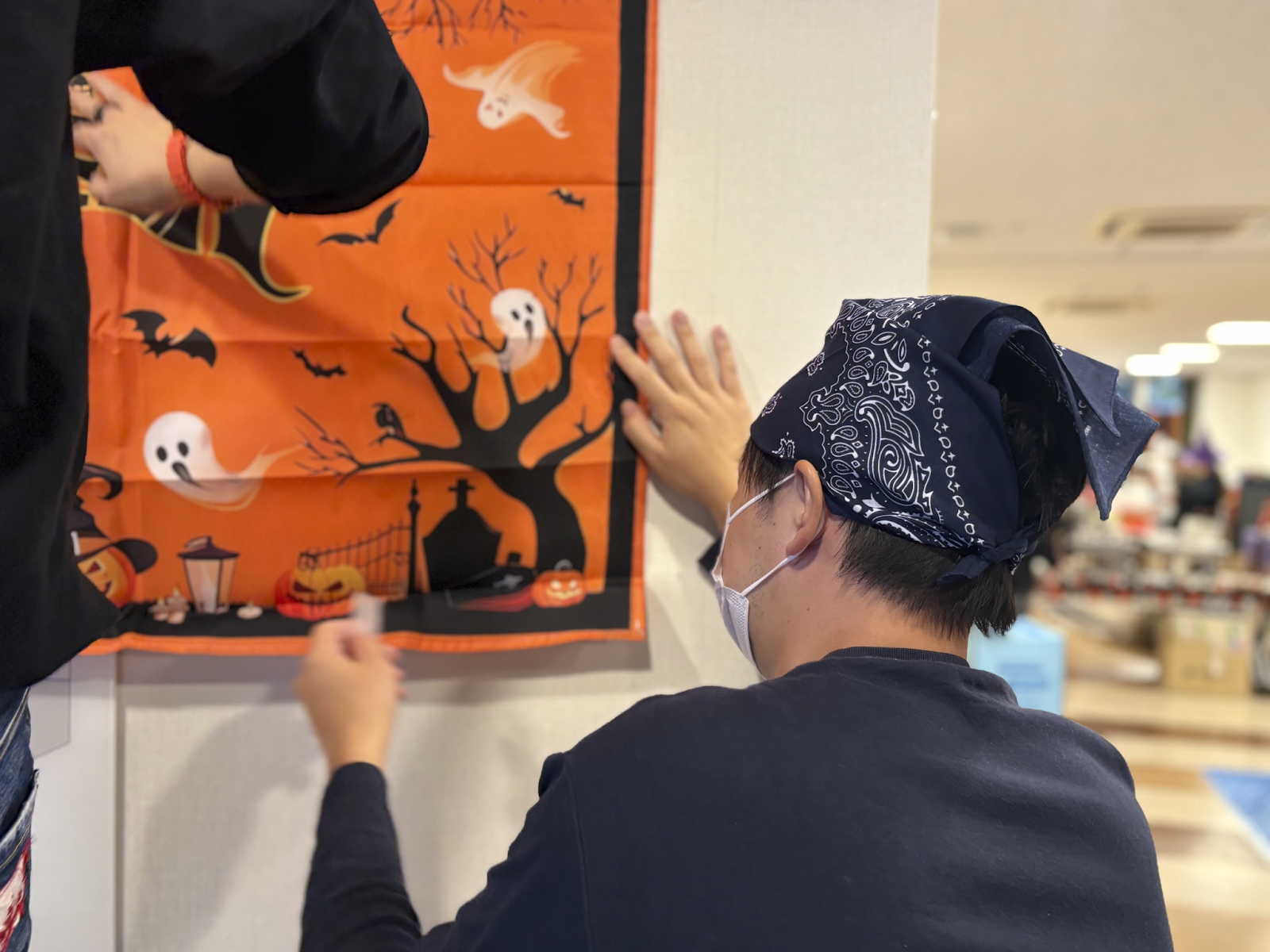 Two people hang an orange Halloween banner depicting ghosts, bats, and pumpkins on a wall.