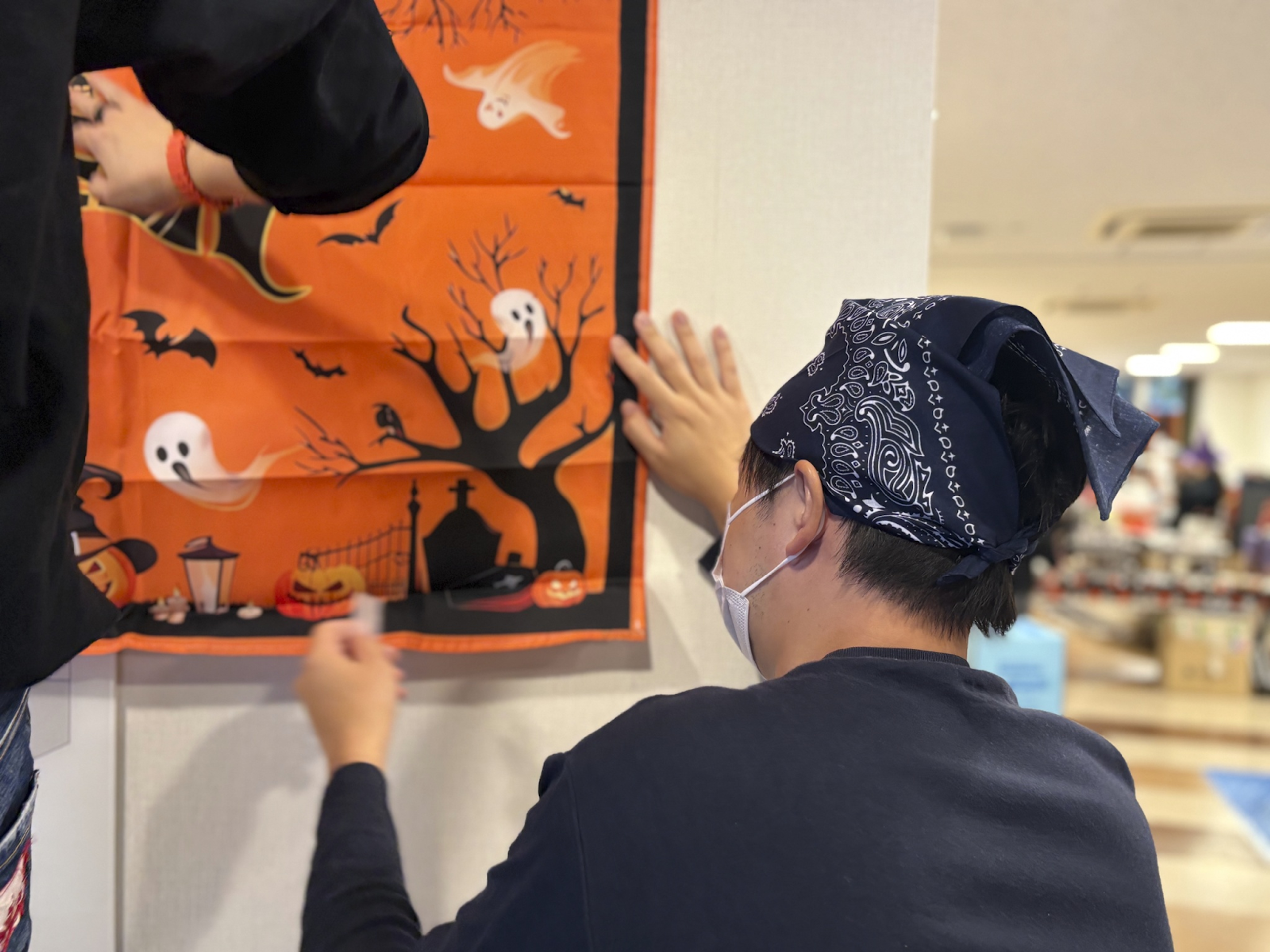 Two people hang an orange Halloween banner depicting ghosts, bats, and pumpkins on a wall.