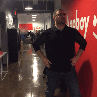 A man stands confidently with his hands on his hips in an office hallway, with a red wall featuring the Appboy logo in the background. Other people are visible in the distance.