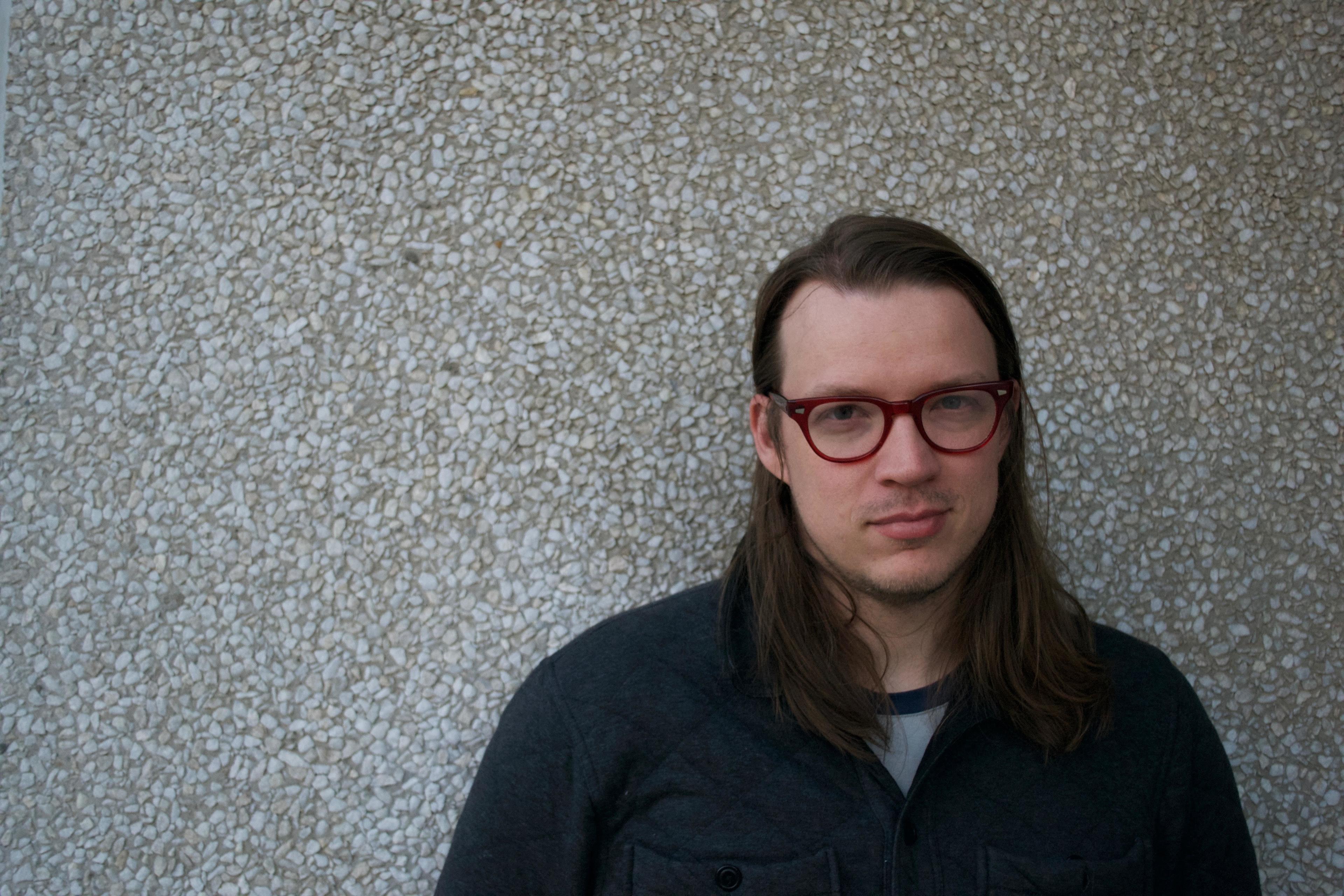 A person with long hair and red glasses stands against a textured gray wall, looking directly at the camera. They are wearing a dark jacket.