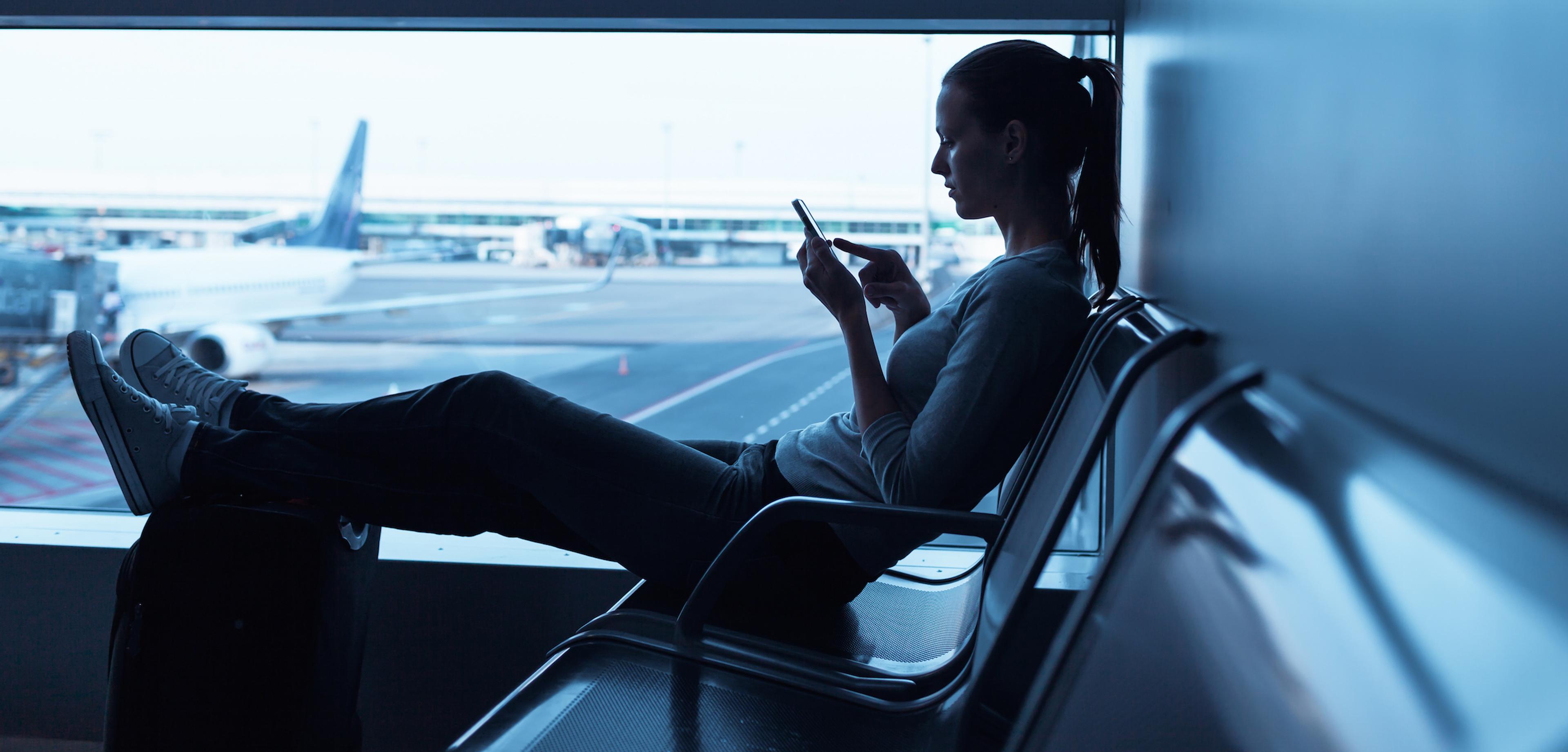 A woman sits in an airport terminal, looking at her phone with her feet resting on a suitcase. Airplanes are visible in the background through the window.