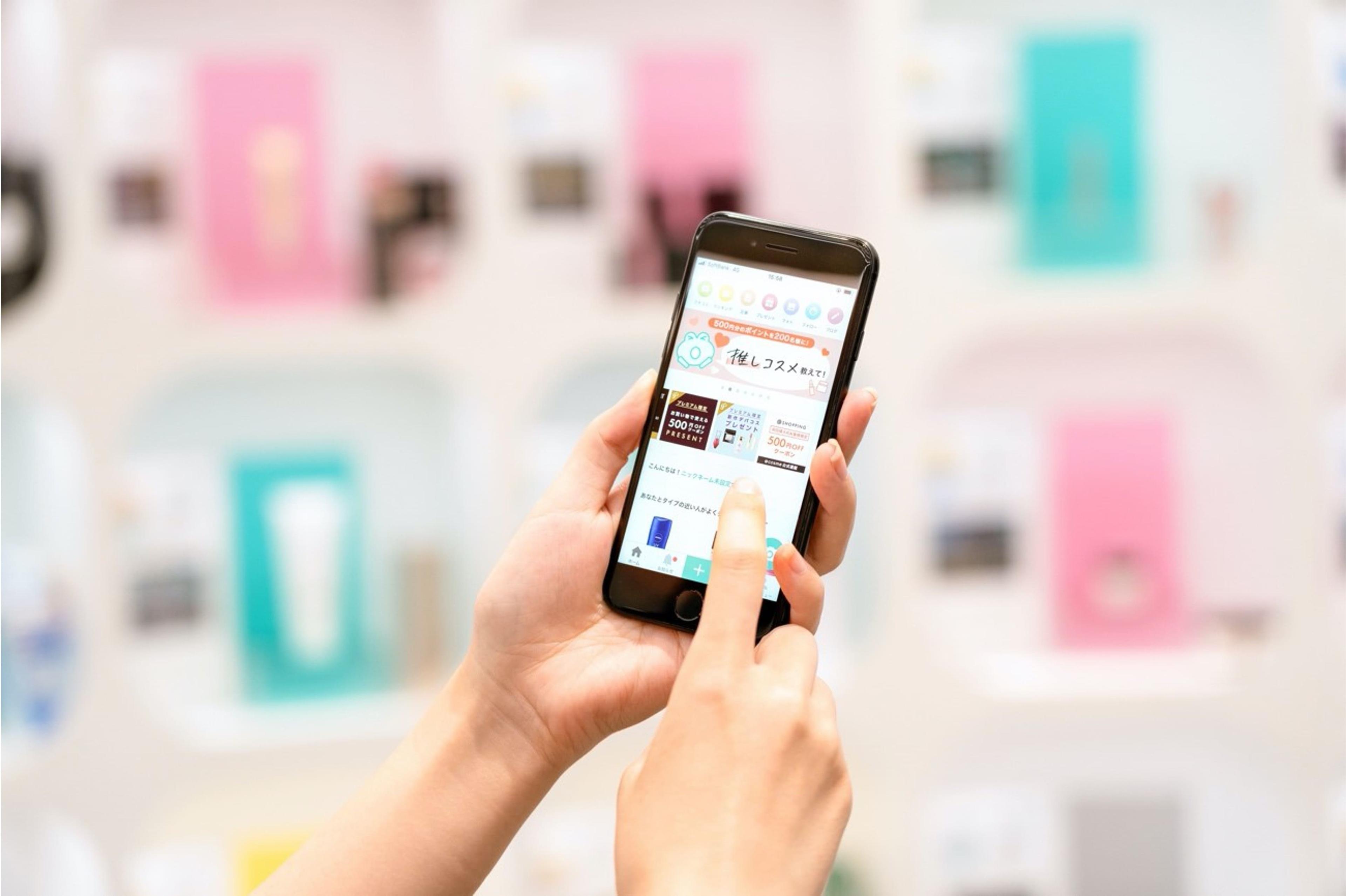 スマートフォンを持つ手が画面をタッチしています。背景にはカラフルな商品が並んでいます。
