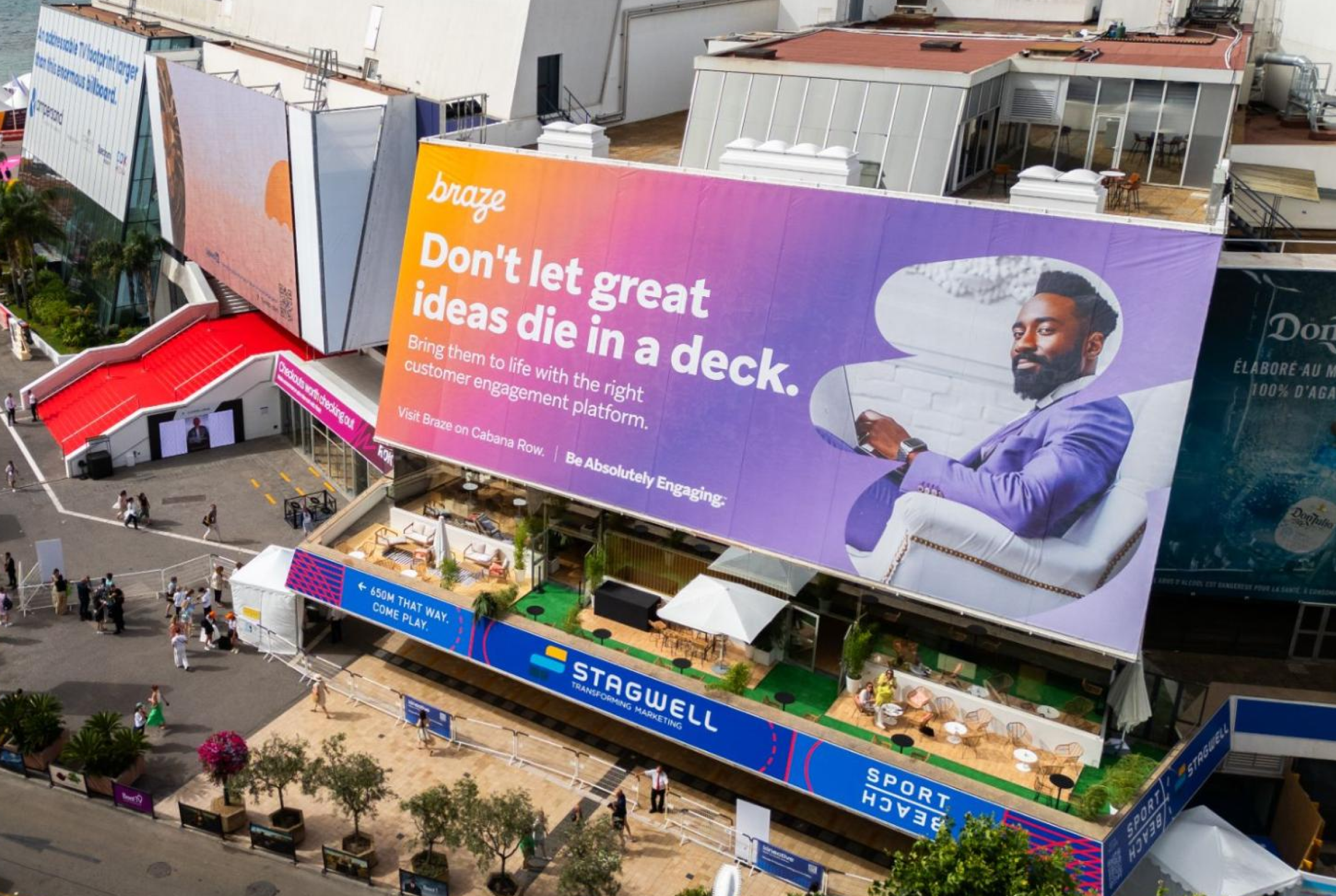 A large billboard displays a promotional message for a customer engagement platform, featuring a man sitting and using a device. The scene includes a vibrant outdoor area with seating and plants below the billboard.