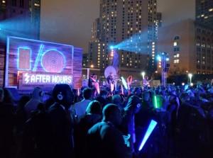 A lively outdoor event at night with a large crowd holding glowing light sticks. A stage features colorful lights and a sign that reads "AFTER HOURS.