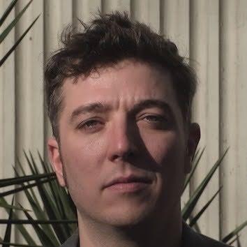 A person with short, tousled hair stands in front of a textured wall, with green plants visible in the background. They have a neutral expression and are looking directly at the camera.