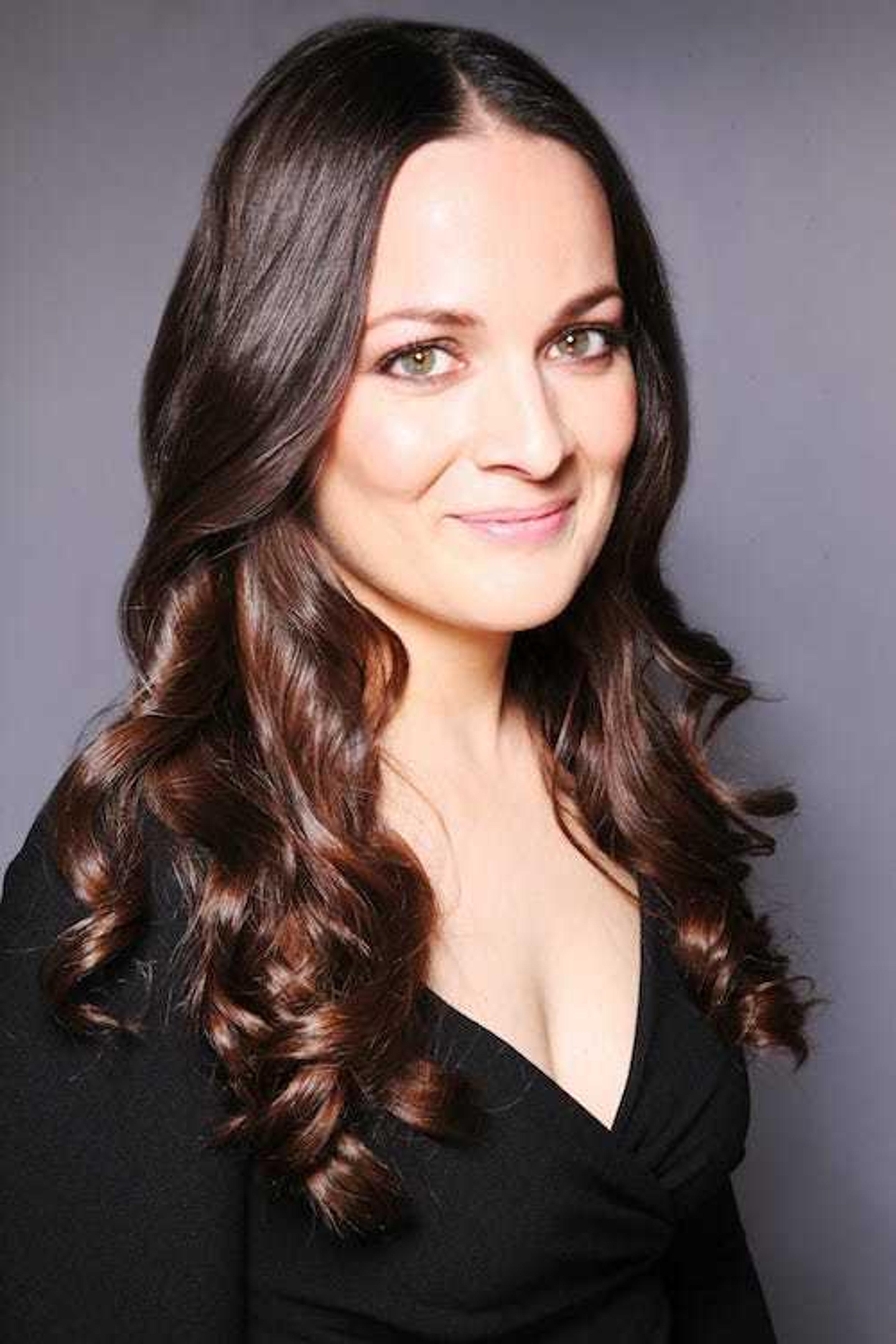 A woman with long, wavy brown hair smiles at the camera, wearing a black top. The background is a soft gray.