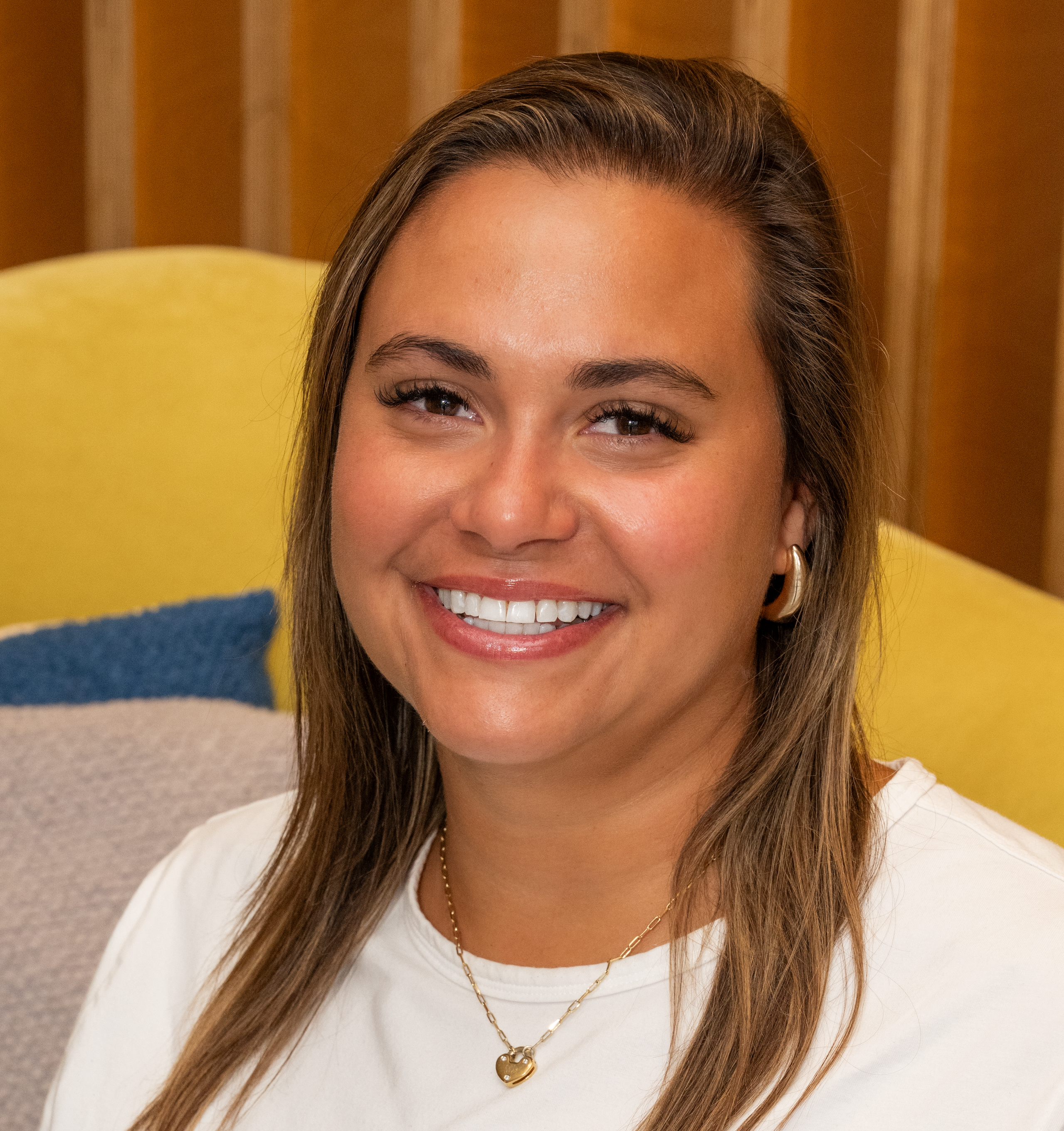 A smiling woman with long brown hair, wearing a white shirt and gold jewelry, sits on a colorful couch. The background features wooden panels and soft cushions.
