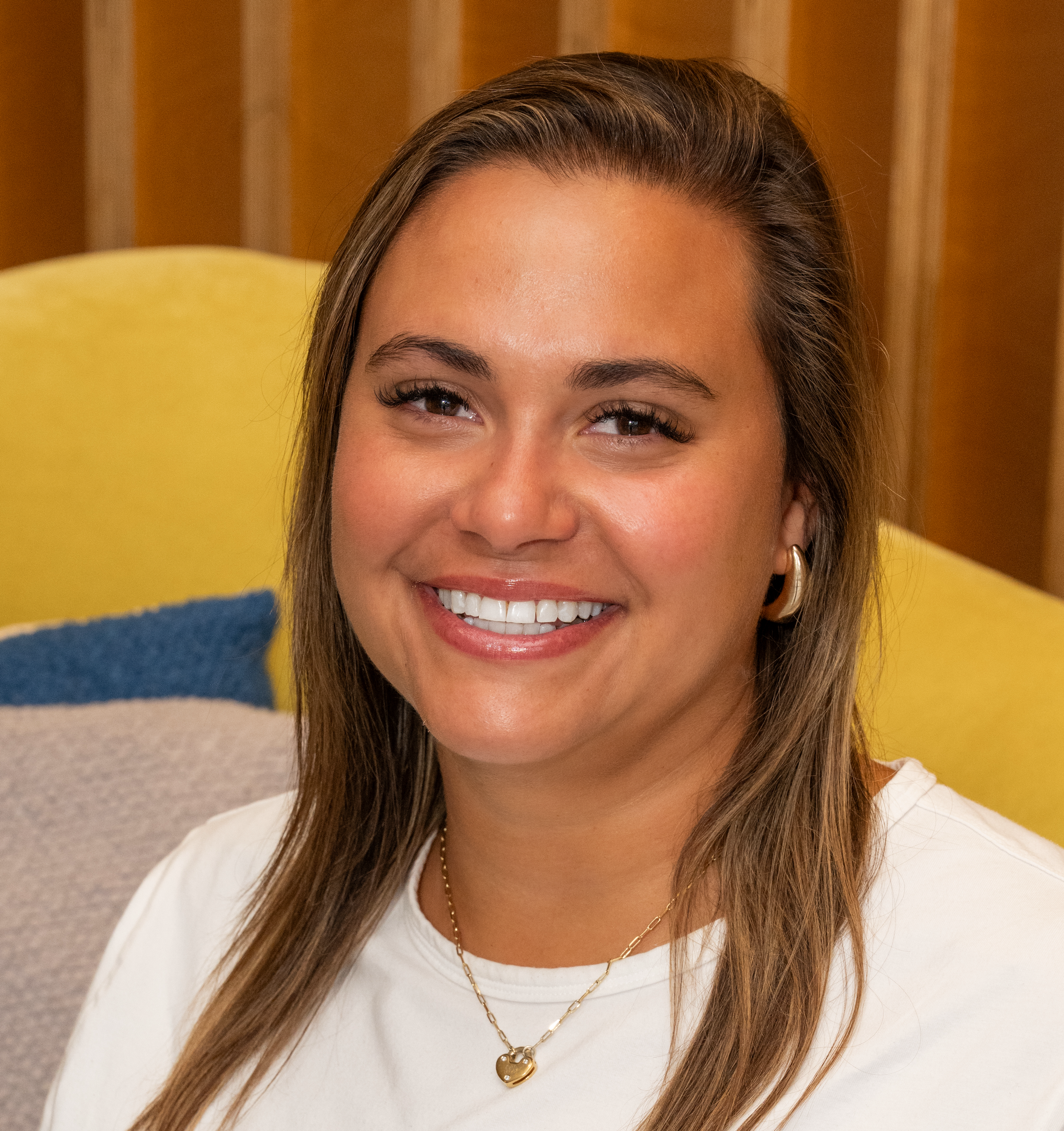 A smiling woman with long brown hair, wearing a white shirt and gold jewelry, sits on a colorful couch. The background features wooden panels and soft cushions.
