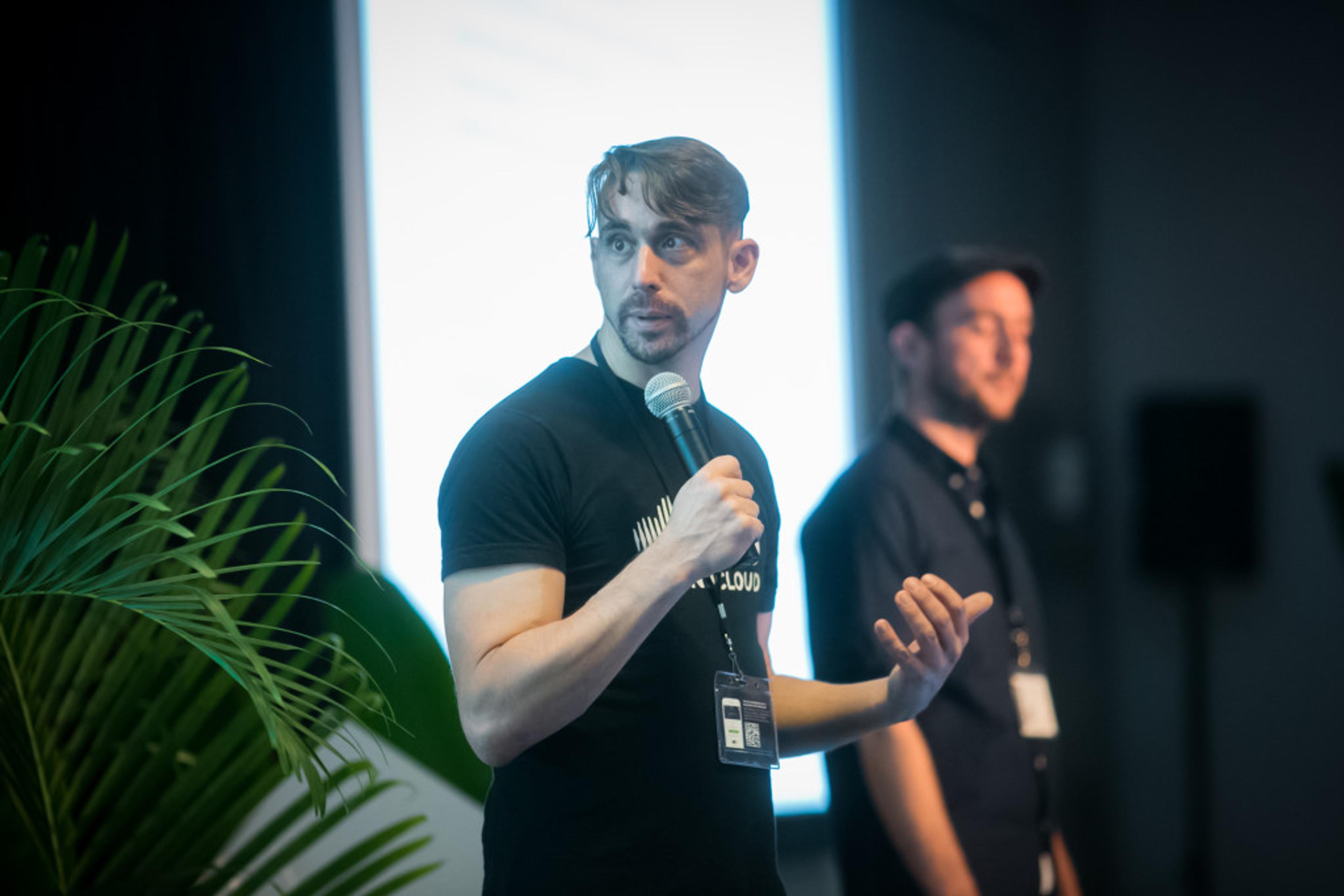 A speaker in a black t-shirt holds a microphone while addressing an audience, with a second person in the background. A plant is visible in the foreground.