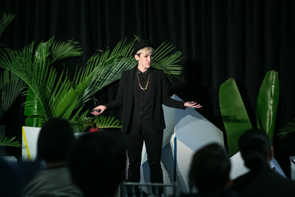 A person in a black outfit stands on stage, gesturing while speaking to an audience. Green plants and geometric shapes are visible in the background.