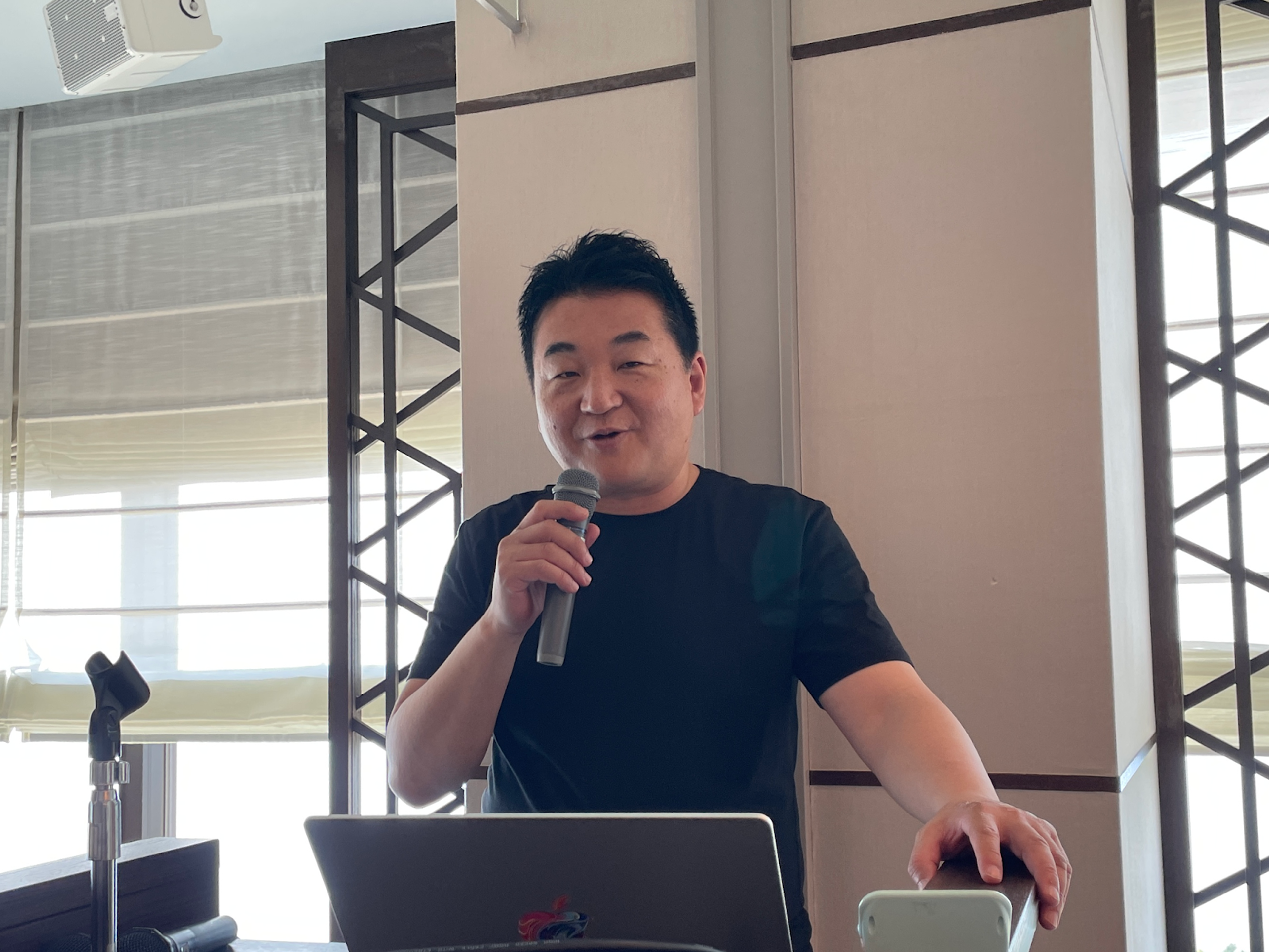 マイクを持って話している男性が、プレゼンテーションを行っています。背景には窓とスライドプロジェクターがあります。