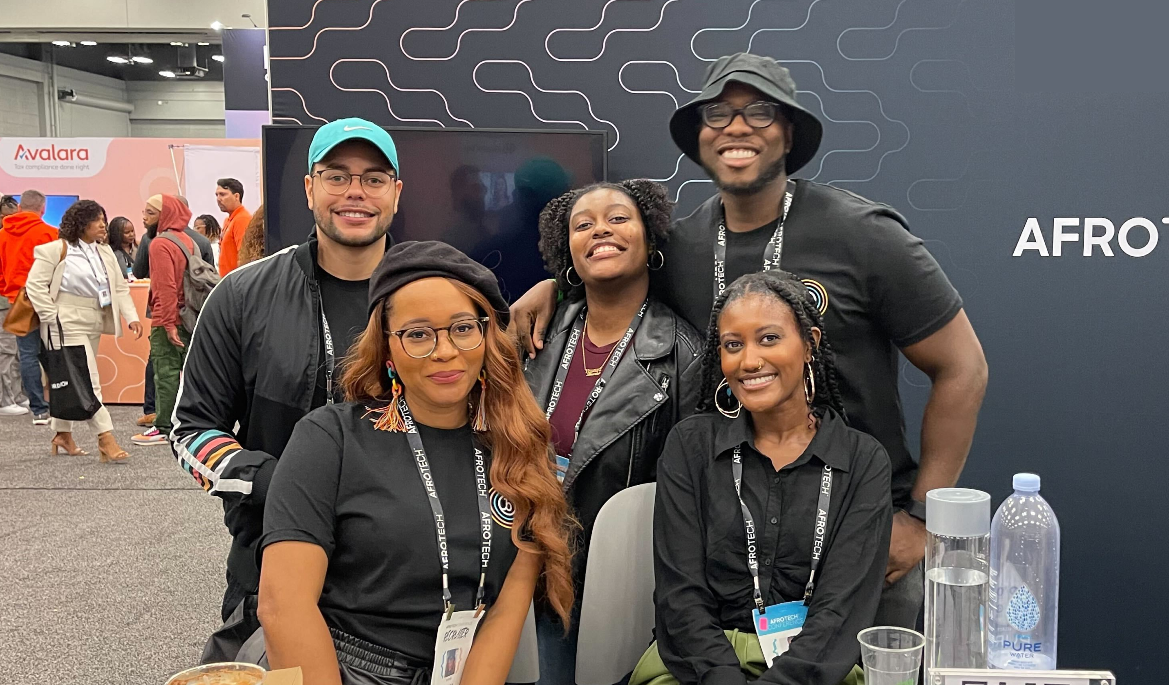 A group of five people poses together at a booth, smiling and wearing matching black shirts. The background features a digital display and other attendees in a conference setting.
