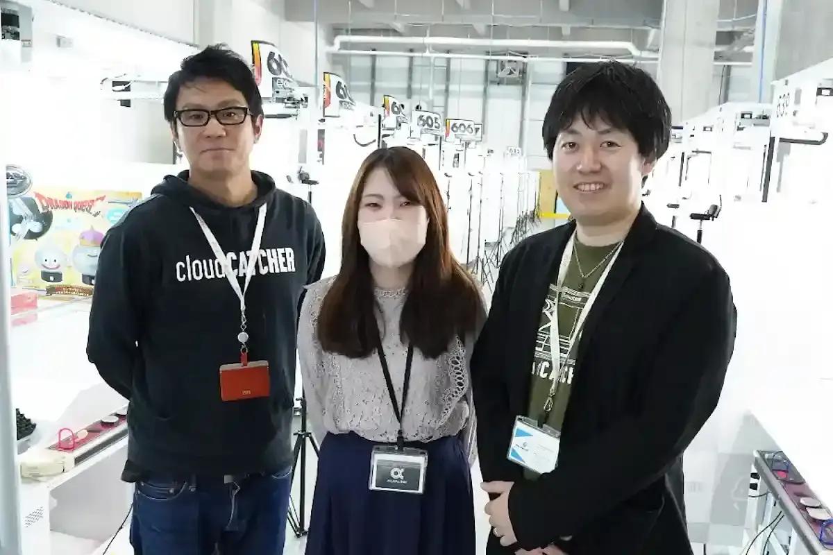 3人の人物が展示会場でカメラに向かって立っています。全員が名札をつけており、背景には展示物が見えます。