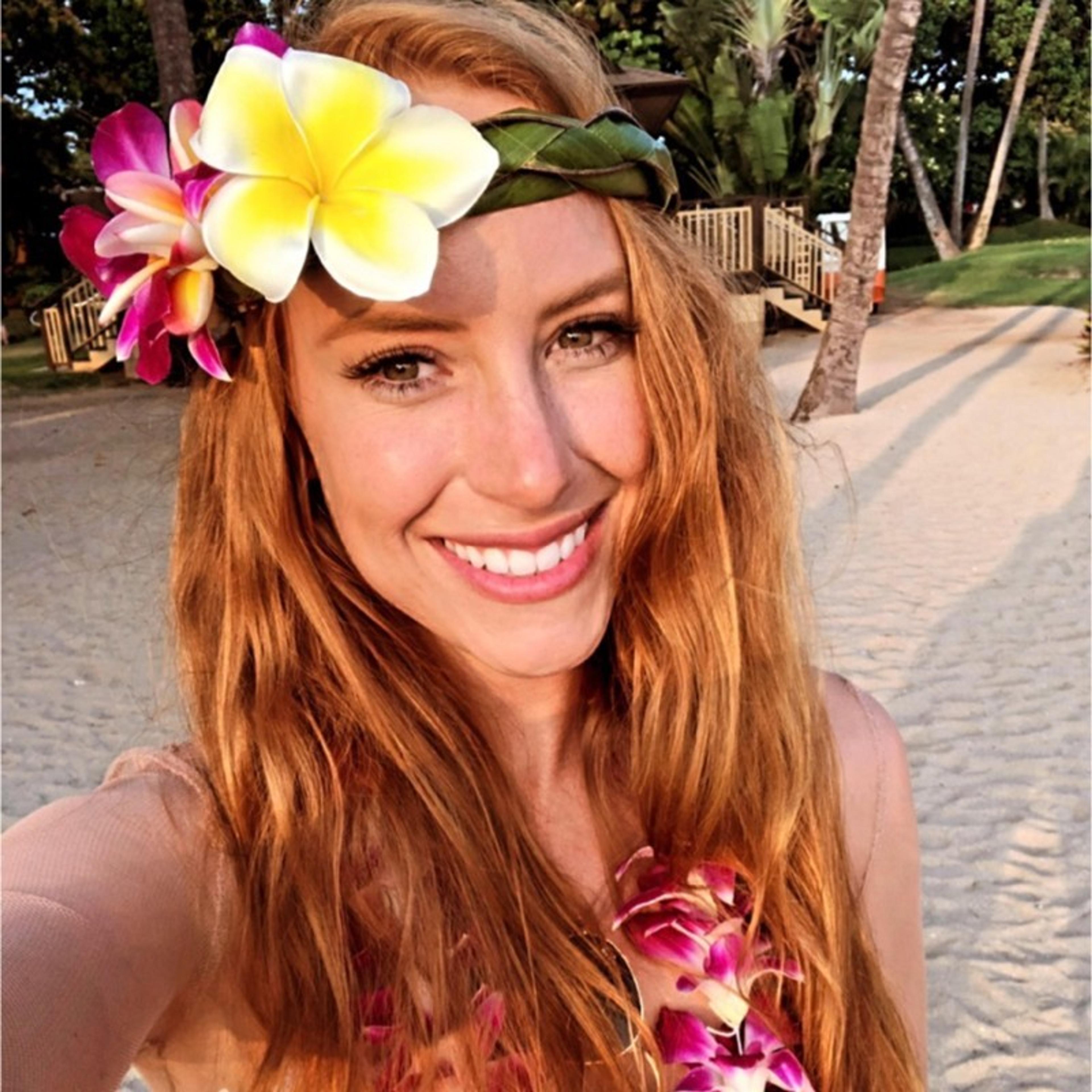 A woman with long red hair smiles while wearing a flower crown and a floral lei on a sandy beach. Palm trees and a wooden deck are visible in the background.