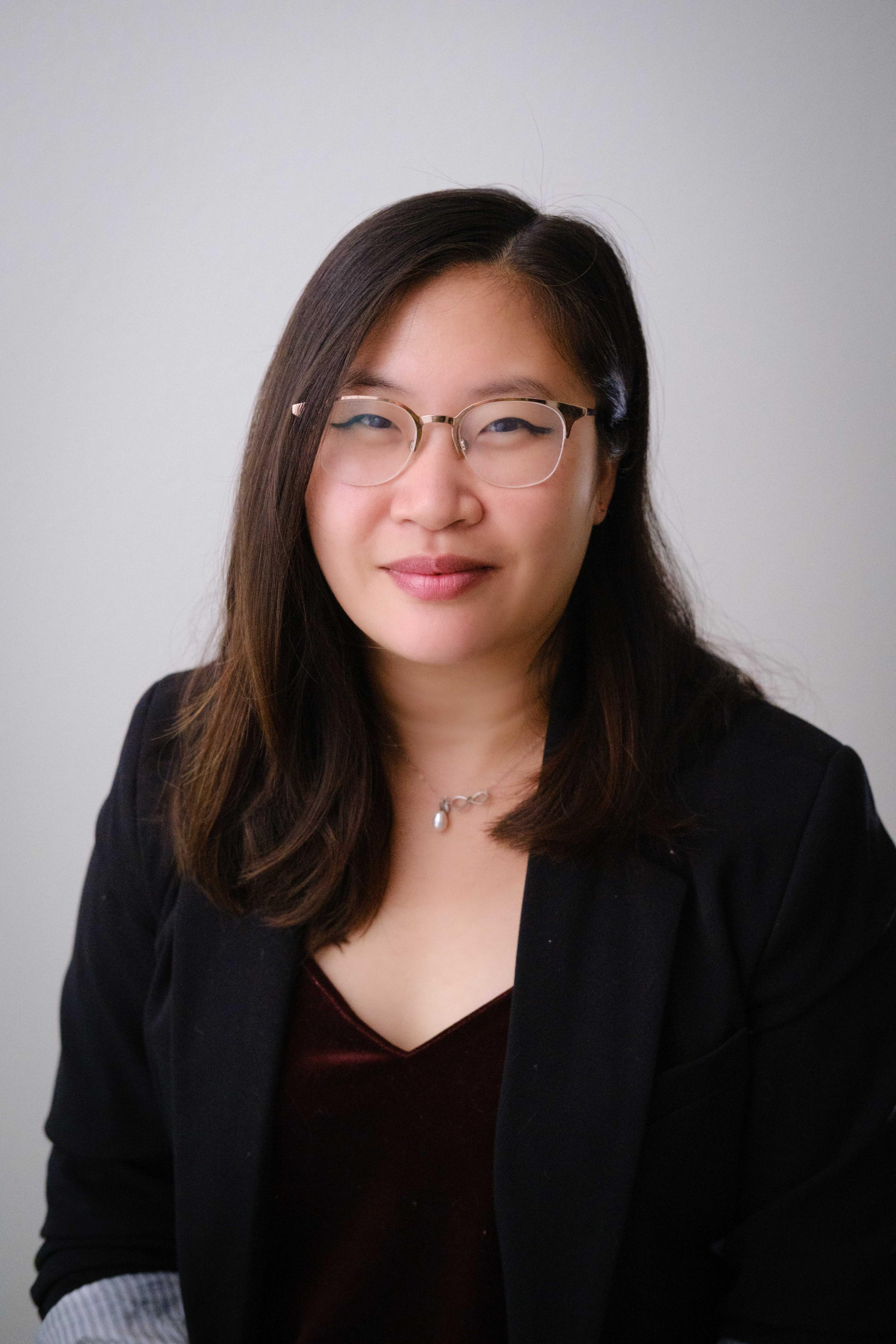A woman with long, dark hair and glasses is smiling while wearing a black blazer over a burgundy top. She is facing the camera against a light background.