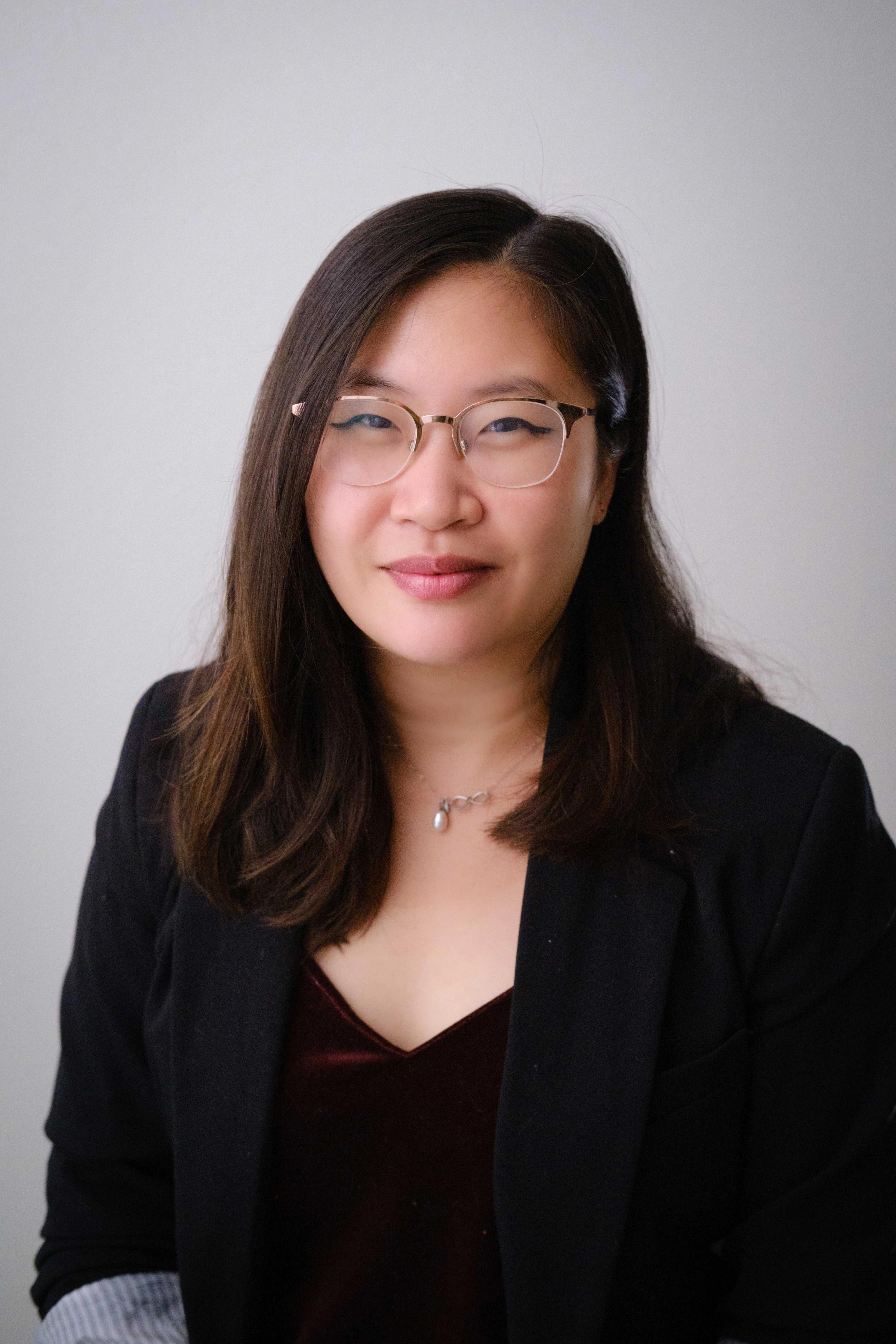 A woman with long, dark hair and glasses is smiling while wearing a black blazer over a burgundy top. She is facing the camera against a light background.
