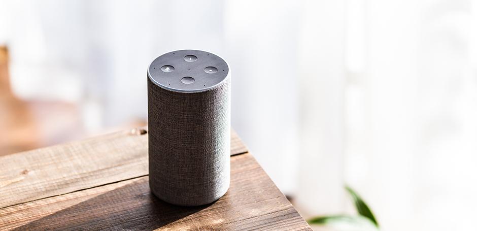 A cylindrical smart speaker with a fabric exterior sits on a wooden table, illuminated by natural light.