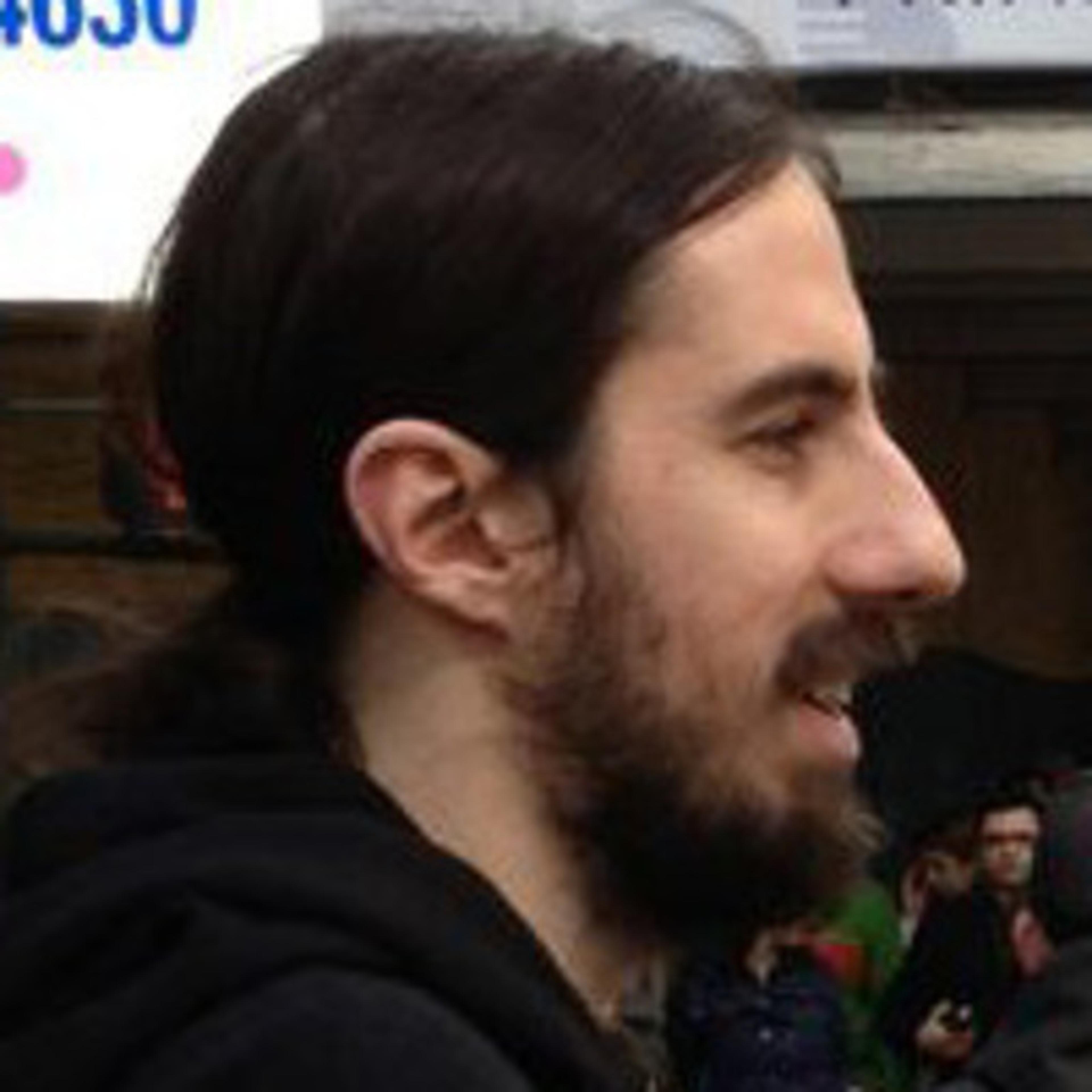 A man with long dark hair and a beard is seen in profile. He is wearing a black hoodie and appears to be outdoors.