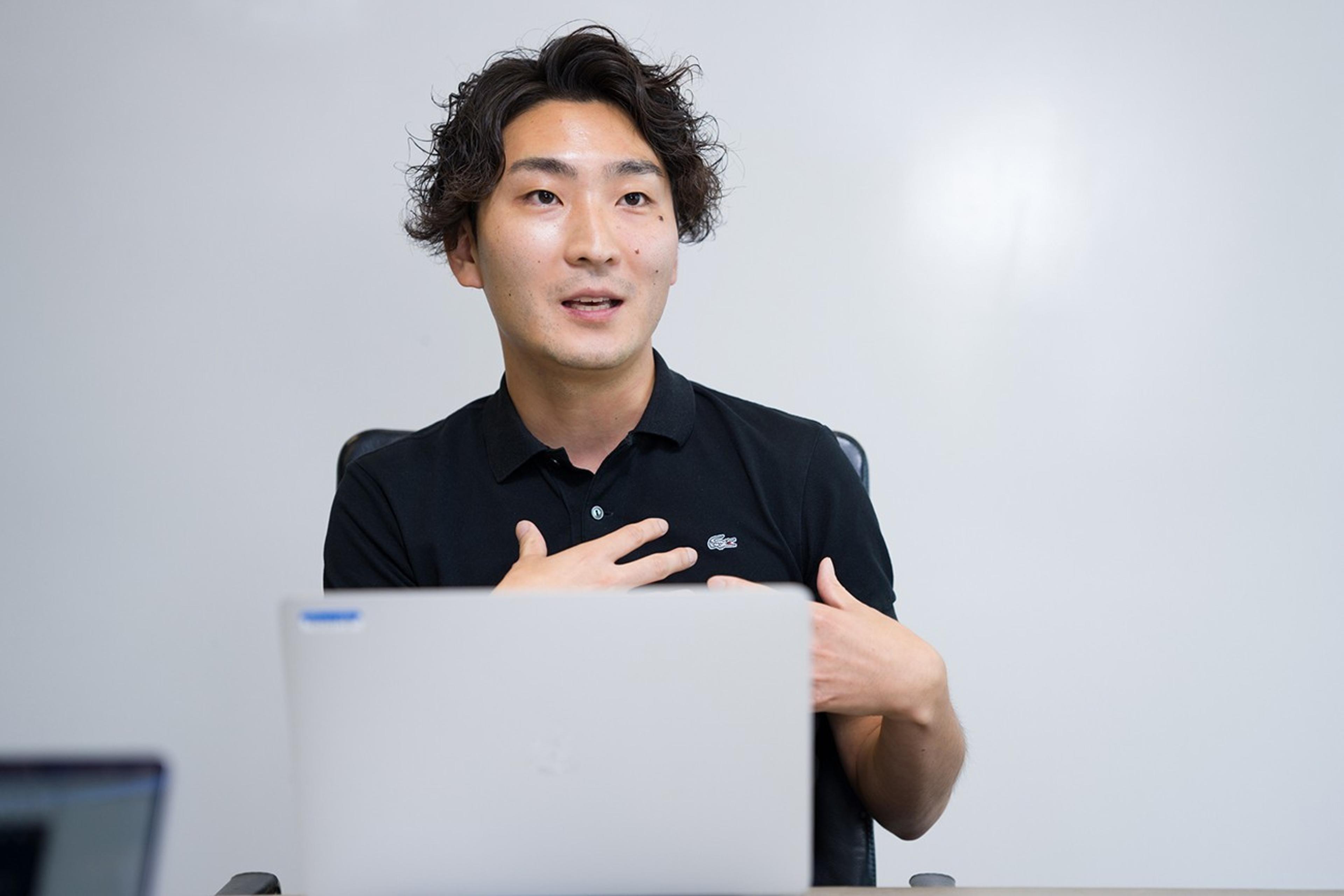 男性が会議室で話している様子。黒いポロシャツを着て、手を使って説明しています。