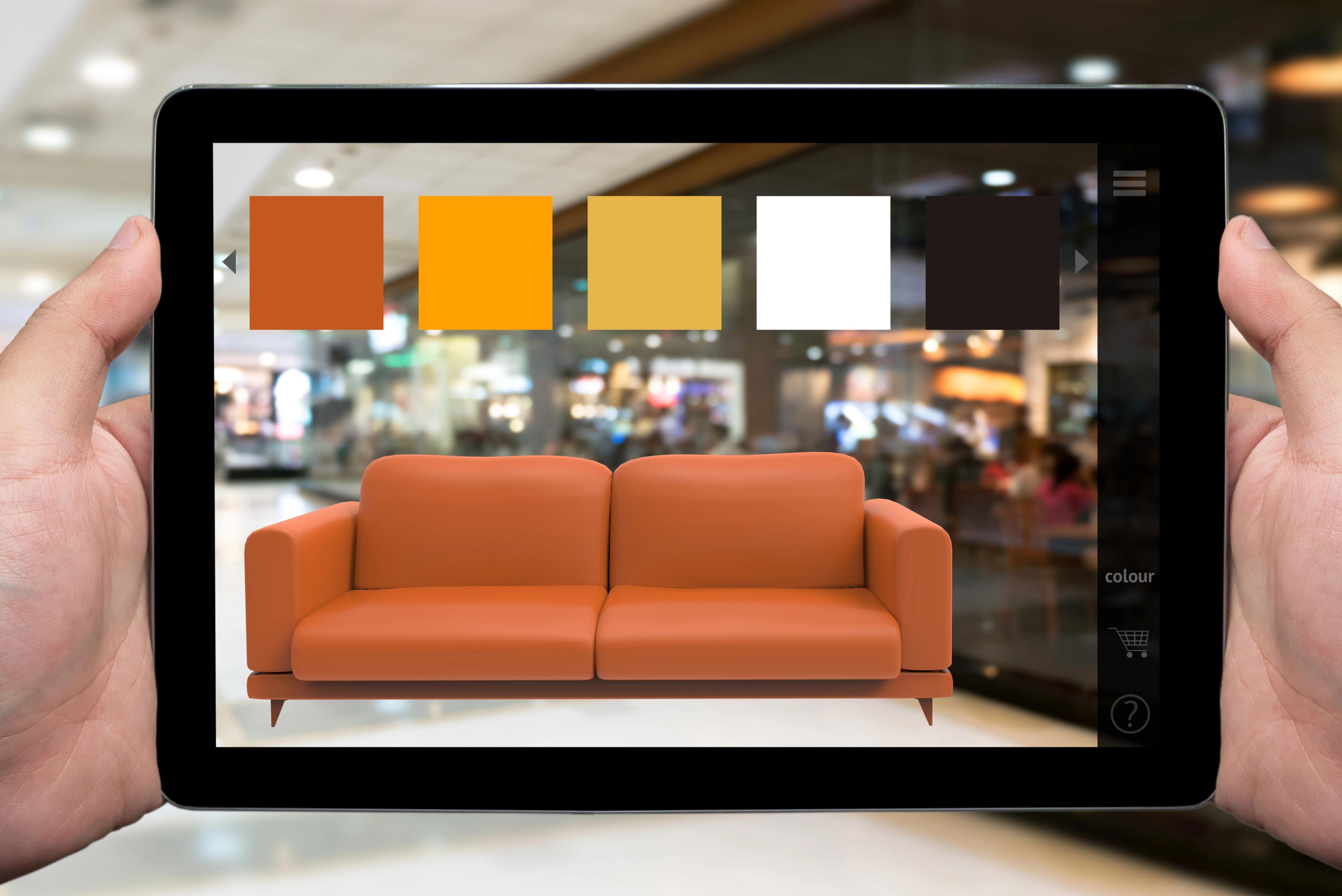 A person holds a tablet displaying a virtual model of an orange sofa with color options above it. The background shows a blurred shopping environment.