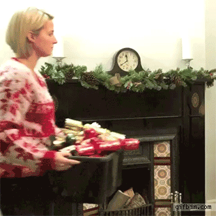A woman in a festive sweater carries a container filled with wrapped gifts towards a decorated fireplace. The scene includes holiday decorations and a clock on the wall.