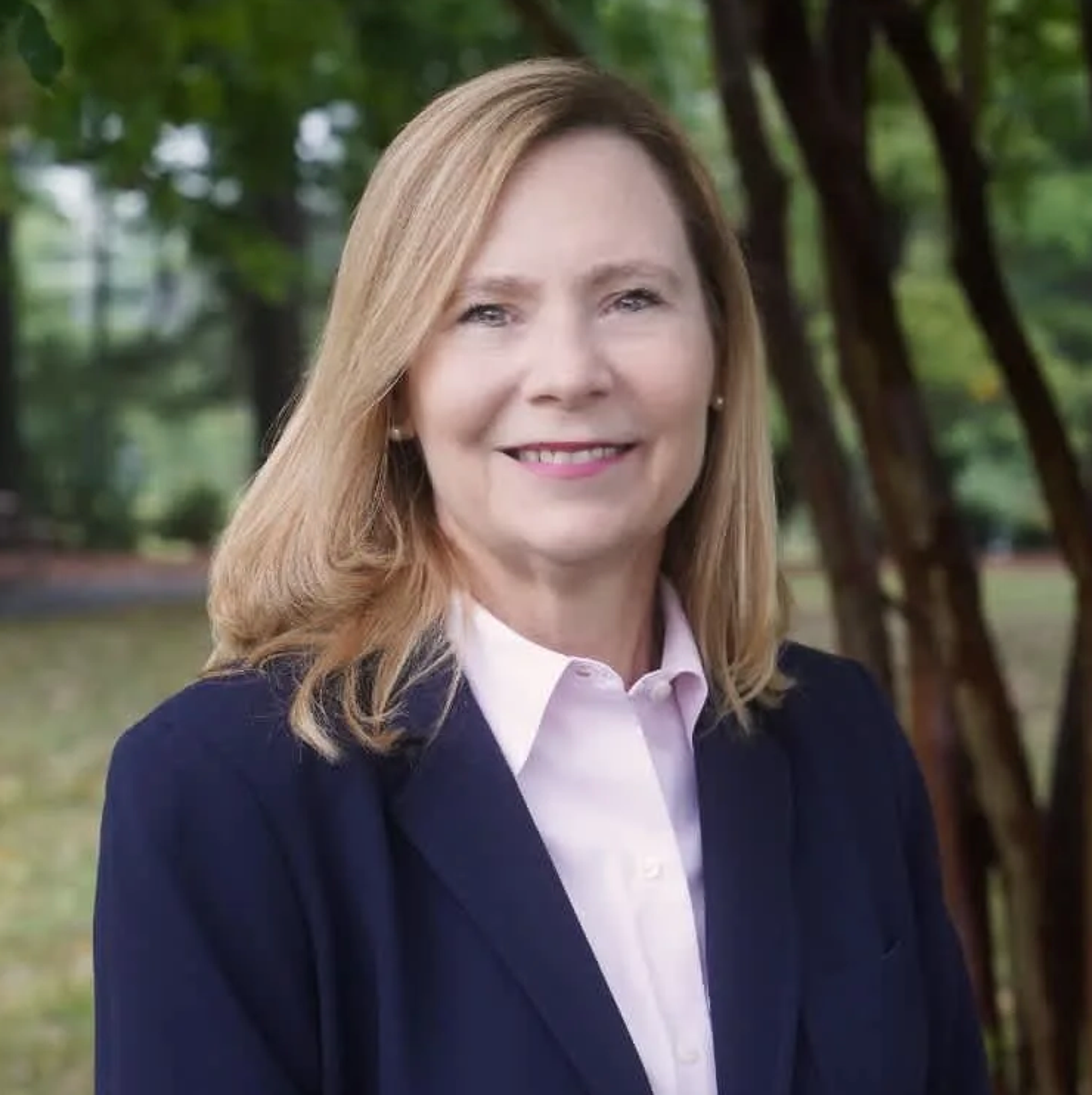 A woman with shoulder-length blonde hair smiles while wearing a navy blazer and light pink shirt, standing outdoors with trees in the background.