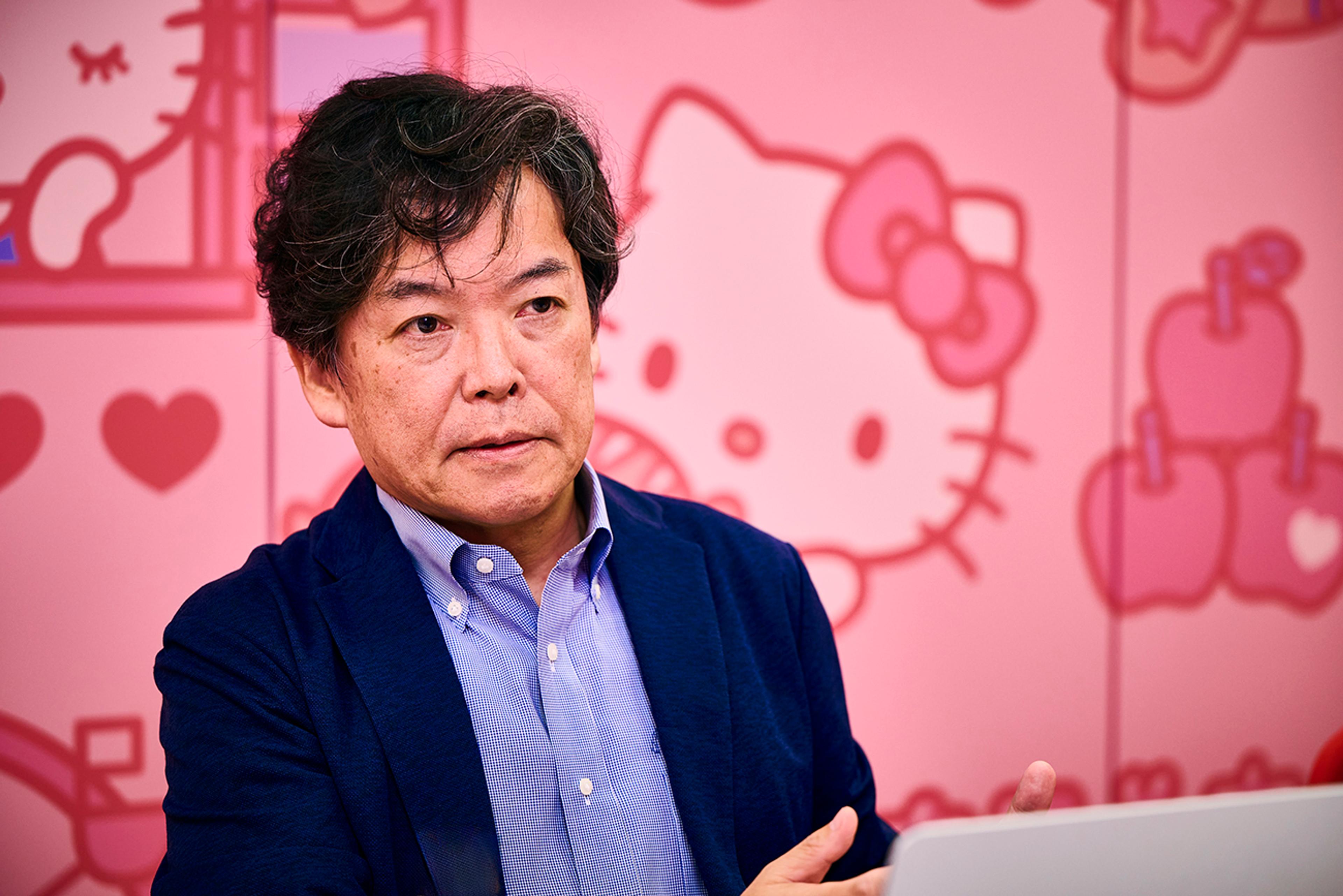 青いシャツとブレザーを着た男性(サンリオ　デジタル事業本部 デジタル事業開発部 CX推進課 顧客体験プラットフォーム戦略推進担当　田口 歩氏)が、ハローキティのシルエットで飾られたピンクの壁の前に座っている。