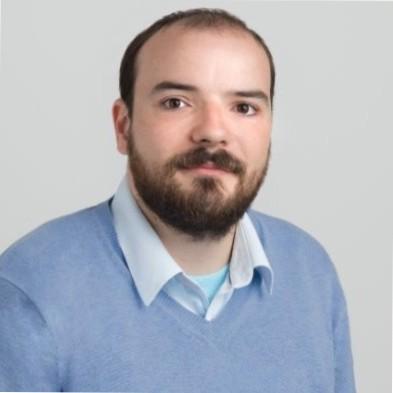 A man with a beard and short hair is wearing a light blue sweater over a collared shirt. He is looking directly at the camera with a neutral expression.