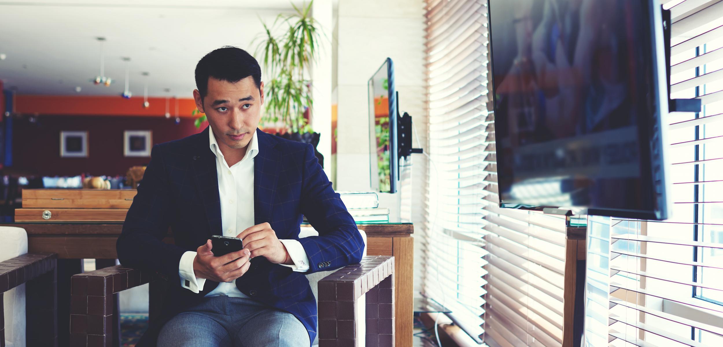 A man in a blazer sits in a modern setting, looking at his phone with a thoughtful expression. Sunlight filters through the blinds, illuminating the space.