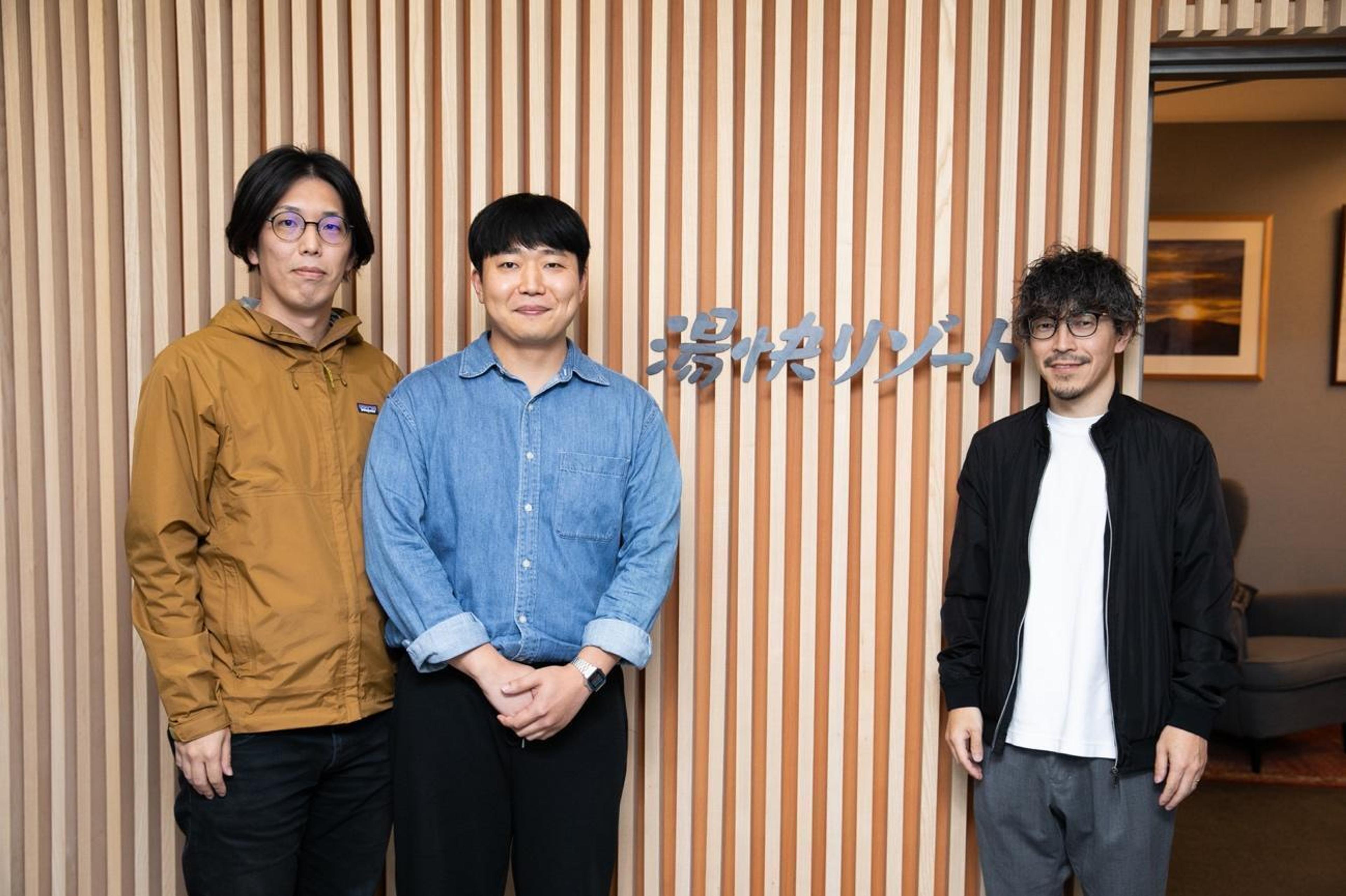 3人の男性が木の壁の前に立っている。壁には「ソリューション」と書かれている。